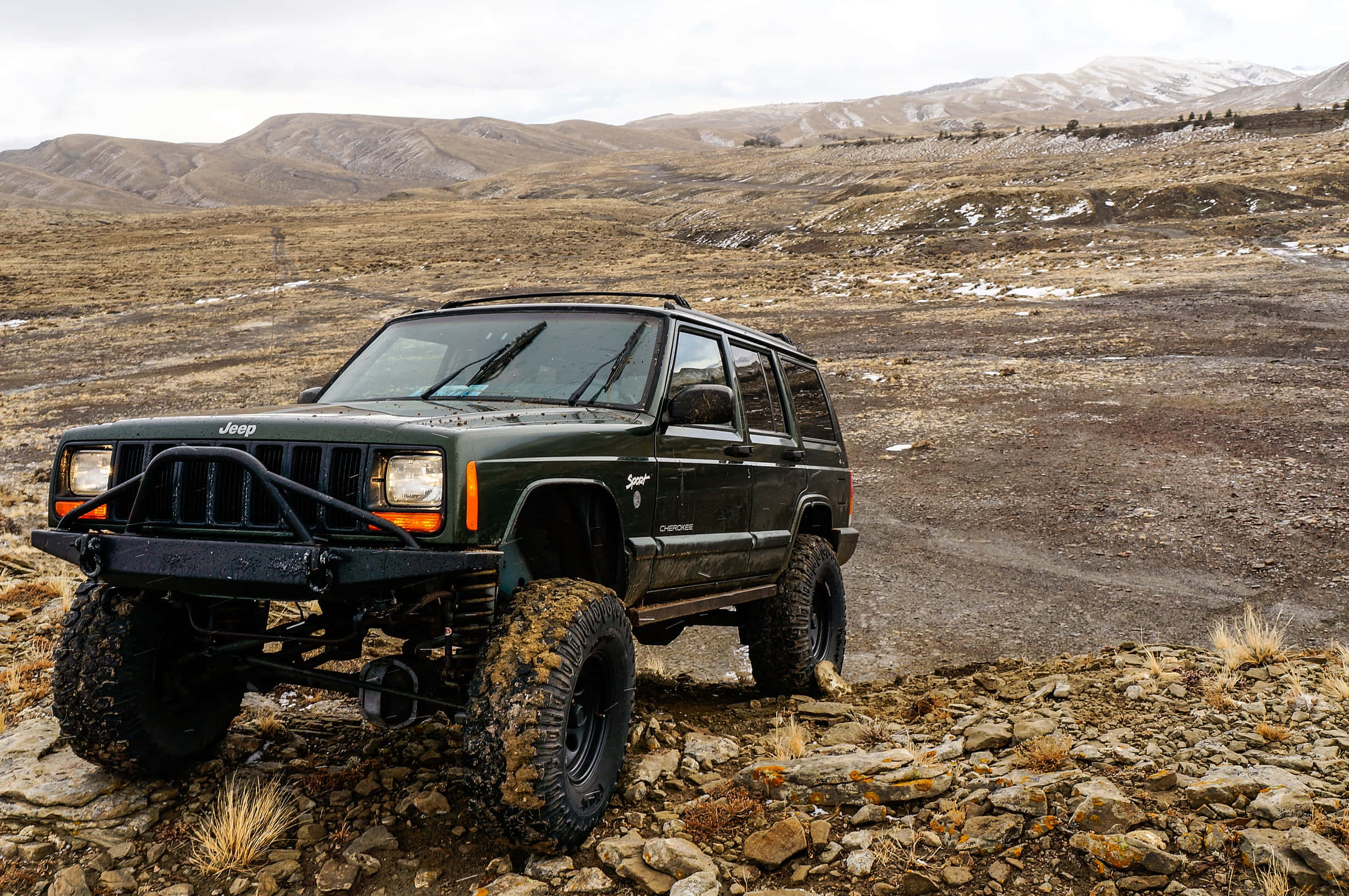 Jeep 動圖 桌布 壁紙 — 一架堅固的綠色Jeep Cherokee配備超大輪胎，在多雲的天空下征服岩石山徑。