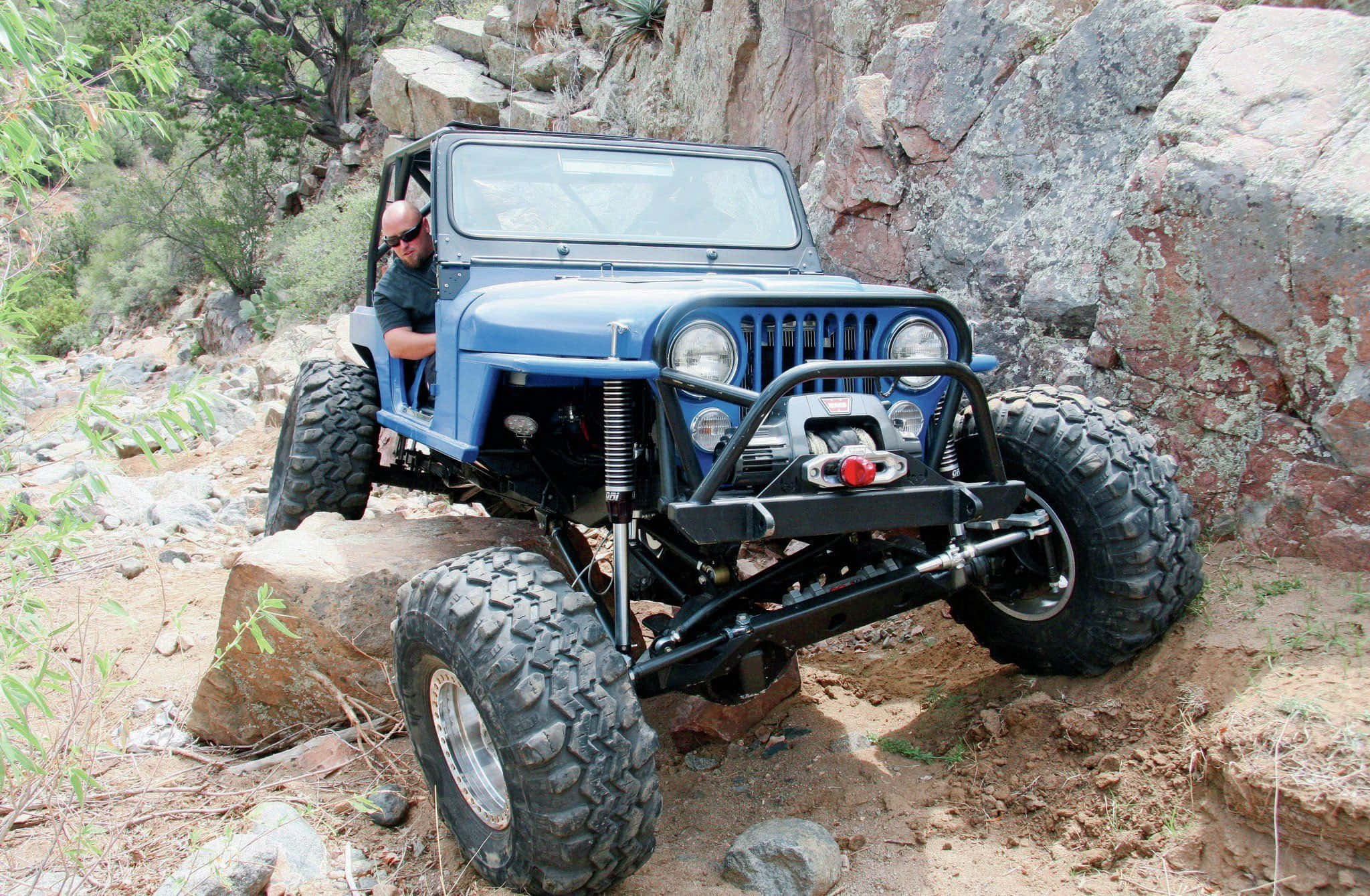 Jeep Cj 桌布 壁紙 — 一架堅固的藍色Jeep Wrangler配備超大輪胎，攀爬陡峭岩石地形，車內司機清晰可見。
