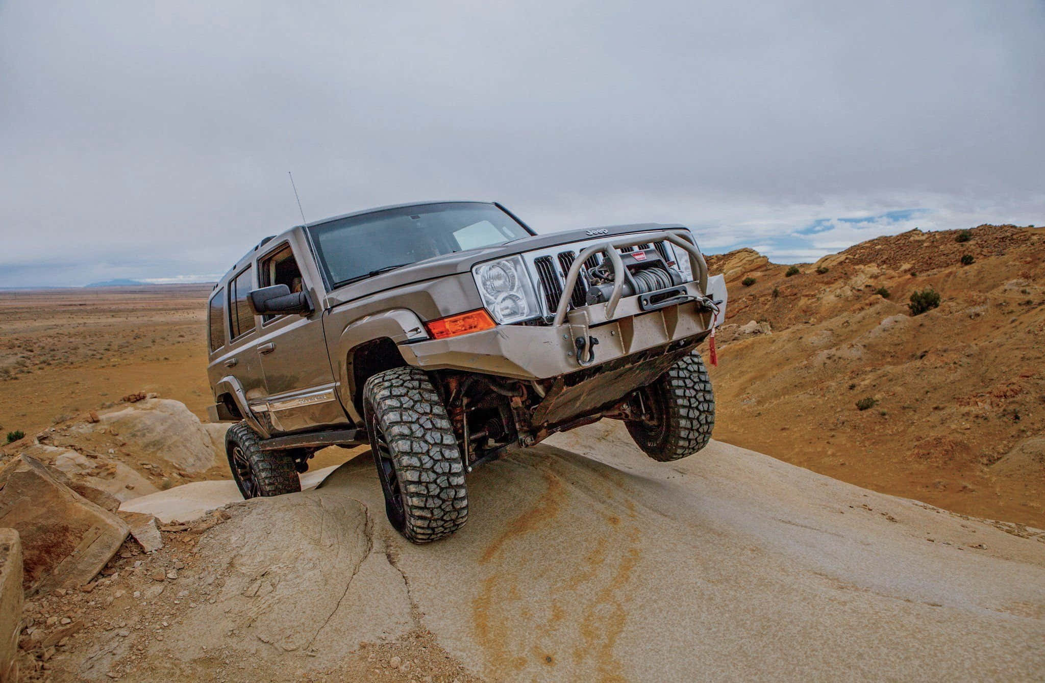 Jeep Commander 桌布 壁紙 — 一架堅固的銀色Jeep Cherokee在陰天下一片廣闊的沙漠中攀爬陡峭的岩石山脊。