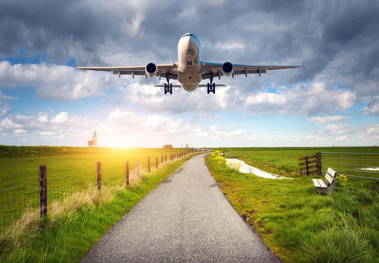 Flight 桌布 壁紙 — 一架大型飛機低飛過一條陽光明媚的鄉村道路，兩旁是綠色田野和木製長椅，天空中有戲劇性的雲層。