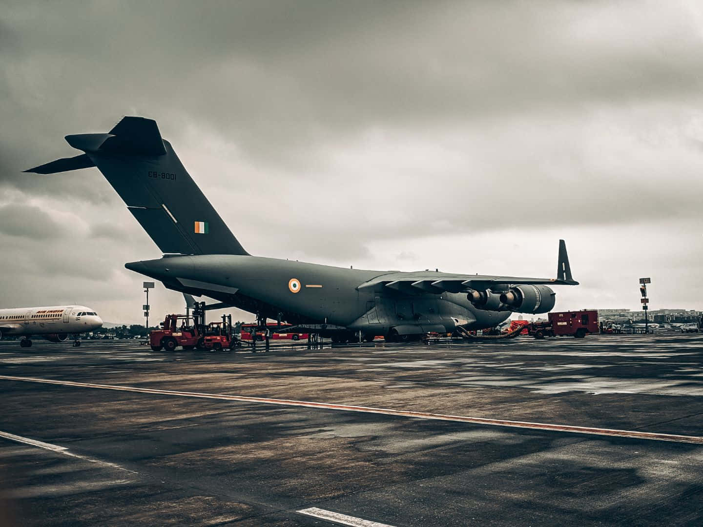 多雲天空下，軍用運輸機在潮濕機場停機坪上，有地面車輛