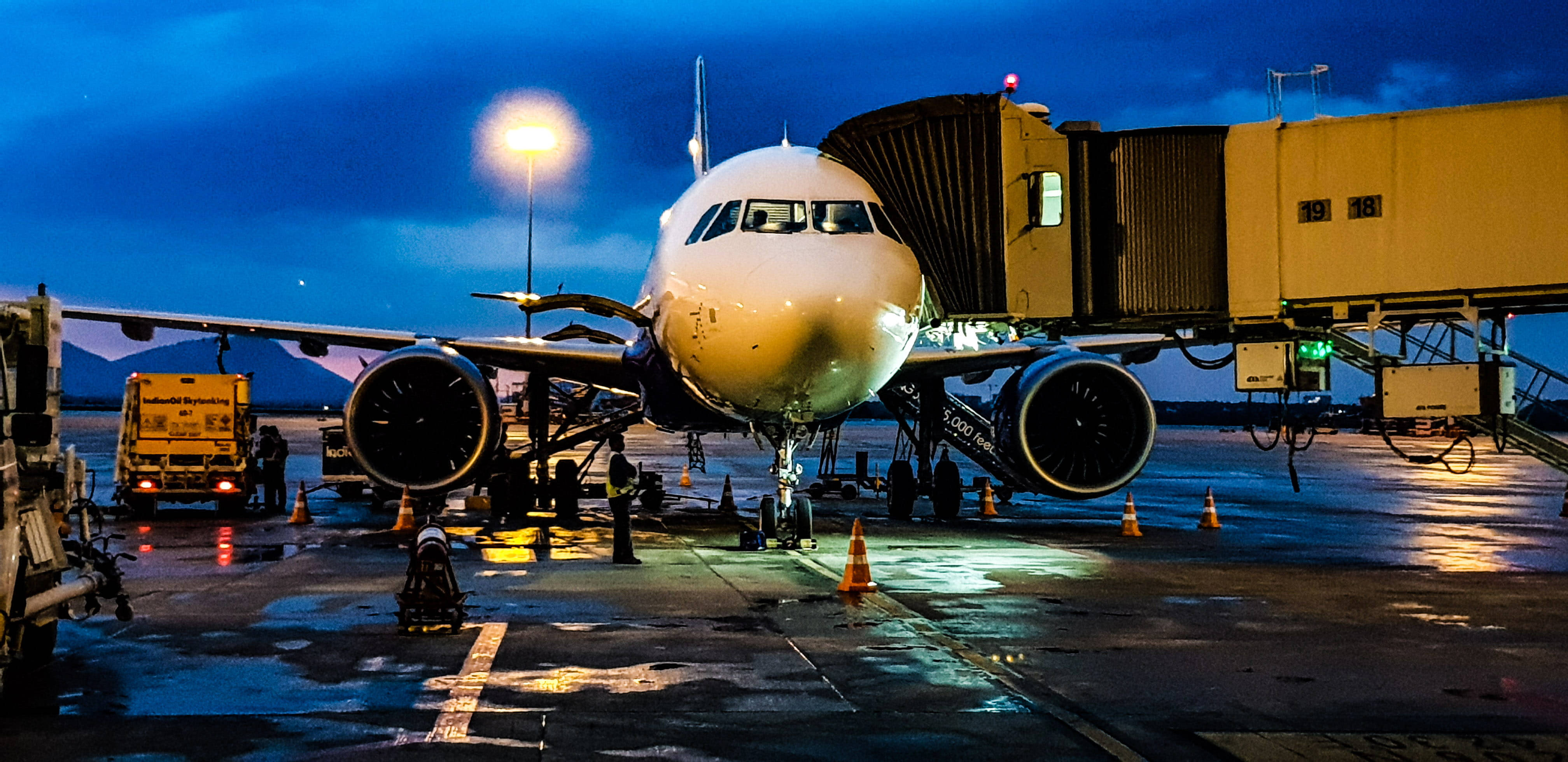 飛機 桌布 壁紙 — 一架大型飛機在暮色天空下停泊在機場閘口，濕潤的停機坪反射出明亮的燈光和遠山。