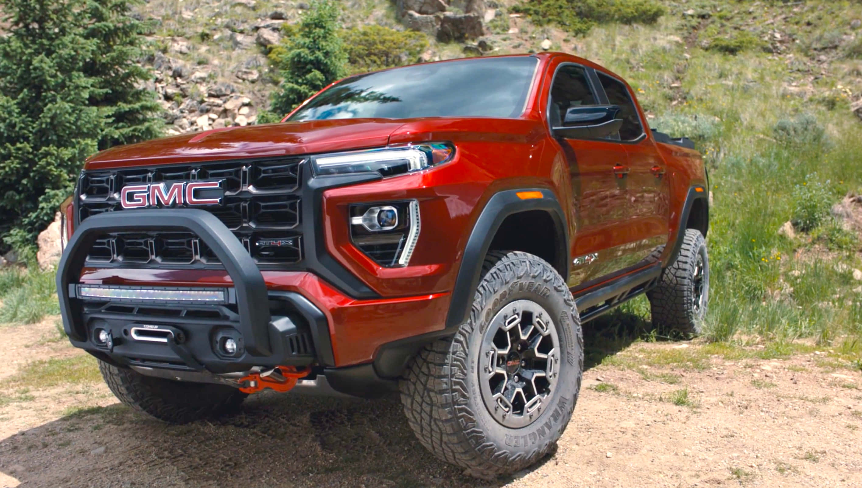 Gmc Canyon 桌布 壁紙 — 一架大膽的紅色GMC Canyon AT4皮卡車，配備黑色越野保險槓和大輪胎，停泊在佈滿綠色灌木的岩石山坡旁邊的泥路上。
