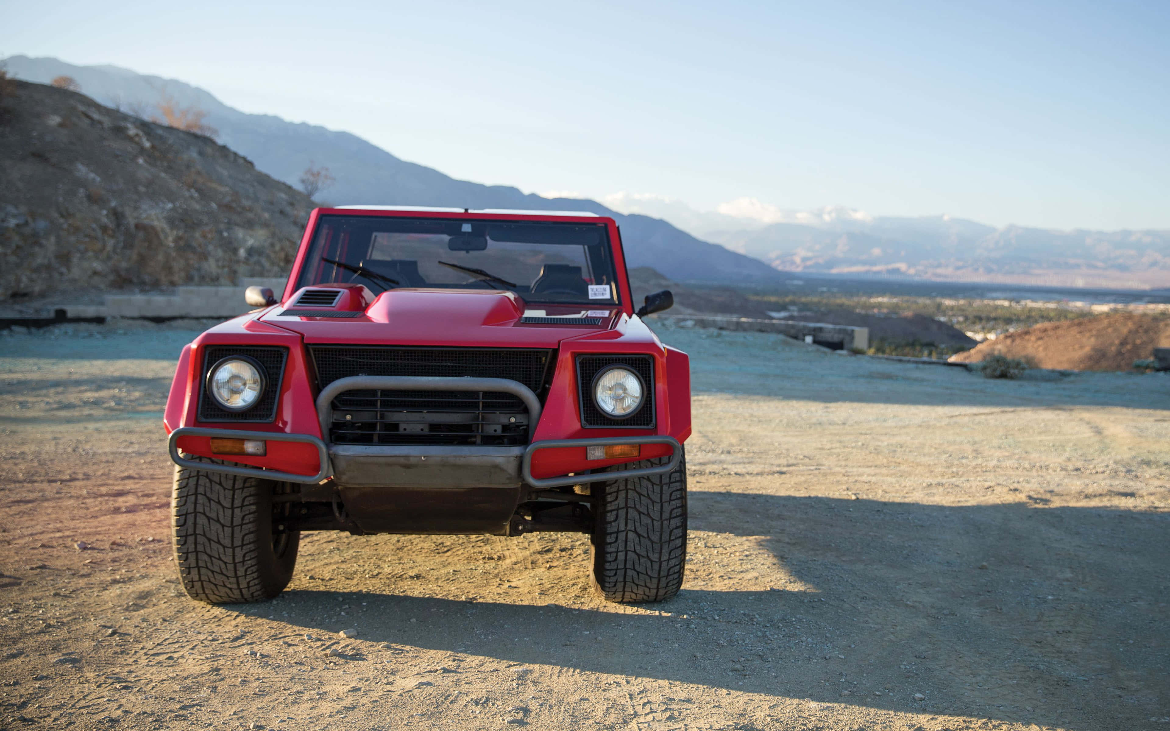 Lamborghini Lm002 桌布 壁紙 — 一架大膽的紅色林寶堅尼LM002正面朝向塵土飛揚的地形，背景是崎嶇的山脈和晴朗的天空。