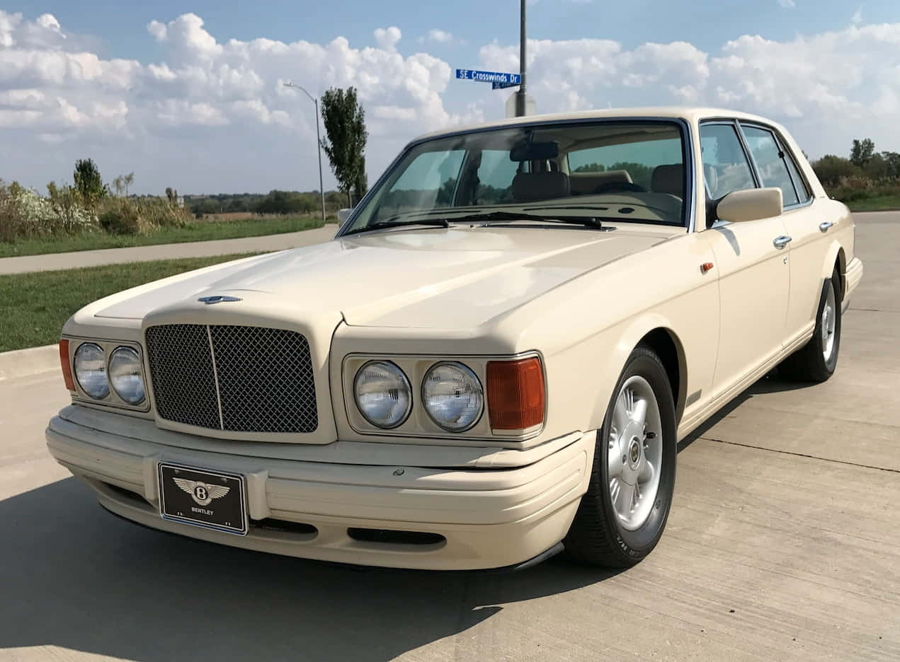 Bentley Eight 桌布 壁紙 — 一架奶油色賓利房車在明亮的日光下閃閃發光，停在混凝土停車場上，背景有藍色路牌和綠化植物。
