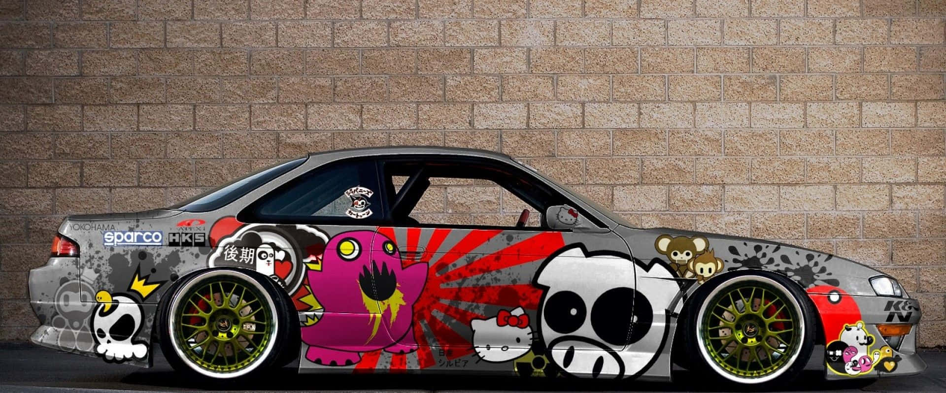 JDM 車 桌布 壁紙 — 一架定制嘅塗鴉覆蓋嘅飄移車，帶有卡通角色同紅色潑濺藝術，停泊喺磚牆前面。