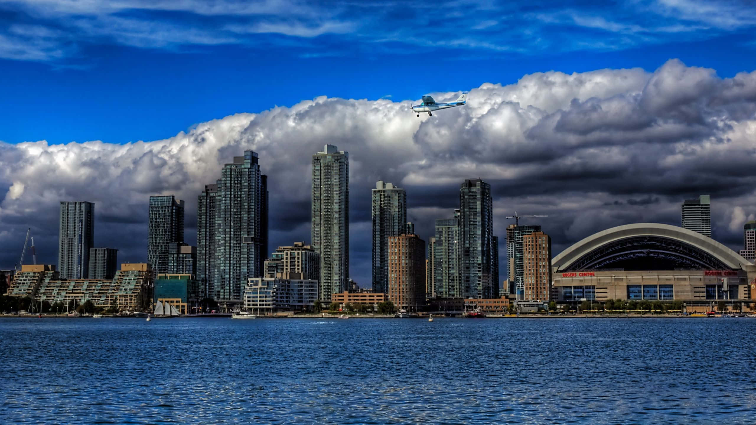Toronto 桌布 壁紙 — 一架小型飛機在藍色水面上倒映的現代城市天際線上方，飛過戲劇性的多雲天空。
