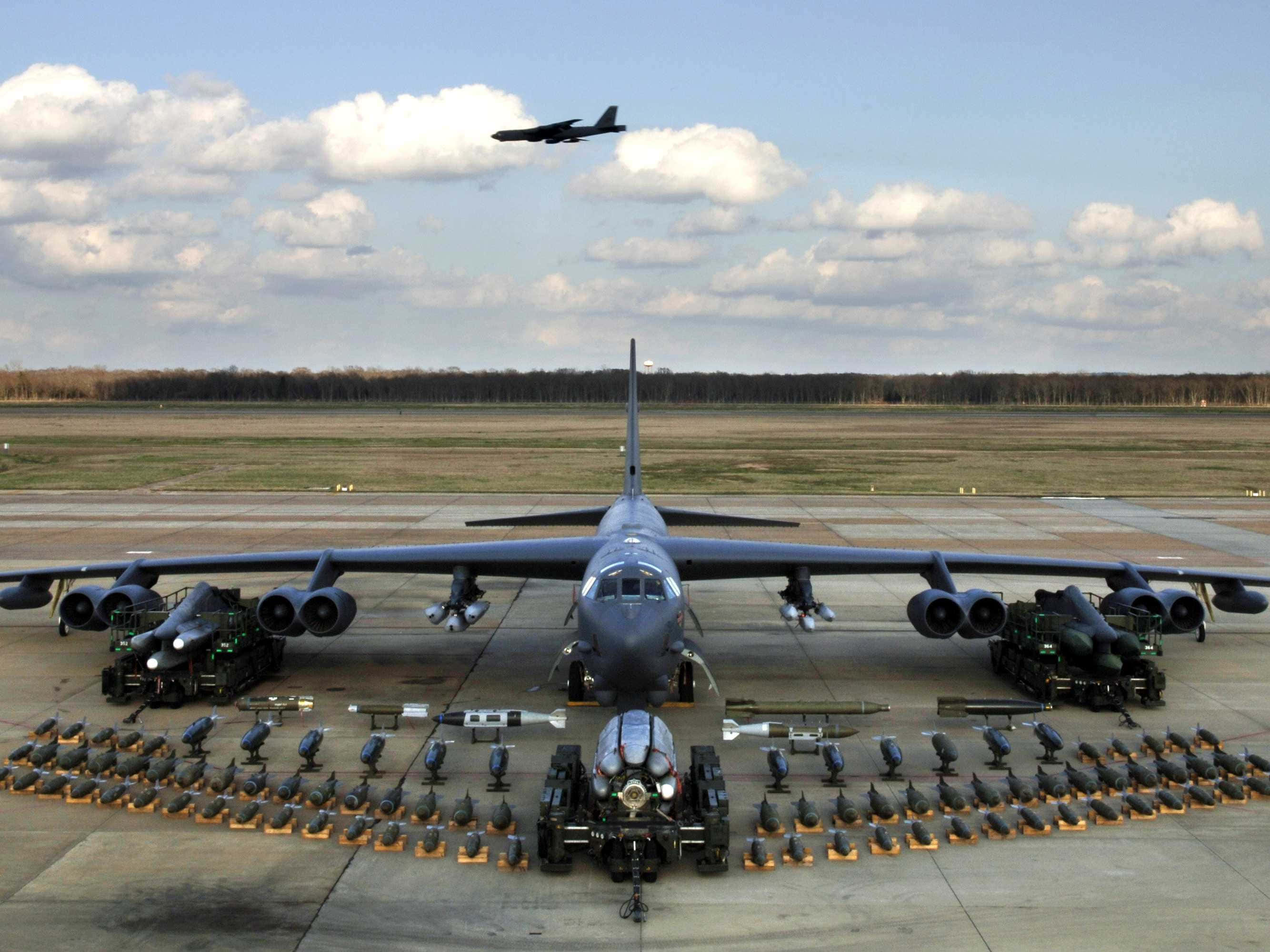 North Korea 桌布 壁紙 — 一架巨大的灰色轟炸機停在機場跑道上，周圍整齊排列著導彈和炸彈，天空部分多雲。