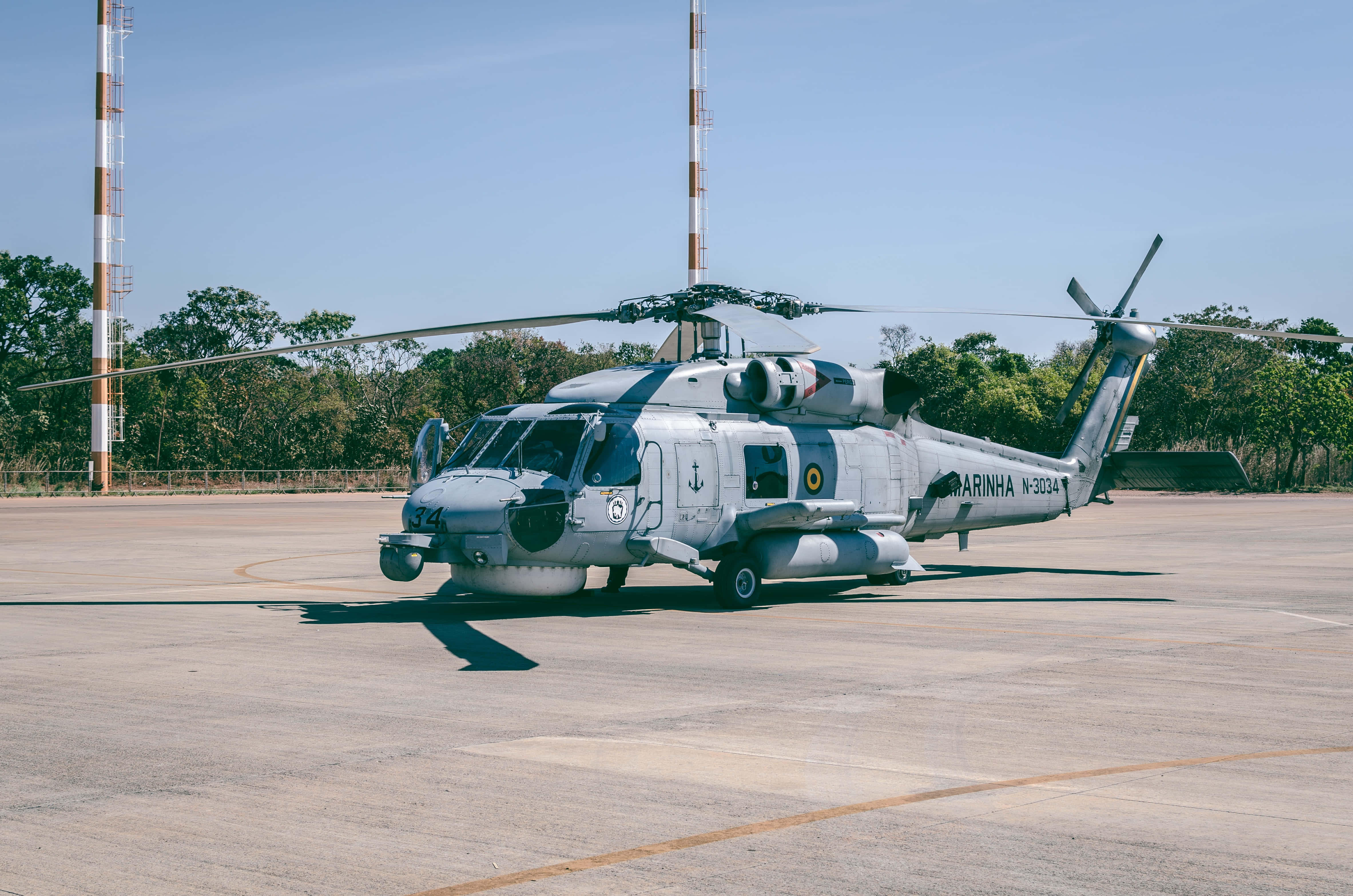 直升機 桌布 壁紙 — 一架帶有 'MARINHA' 標誌的灰色軍用直升機停在停機坪上，背景是晴朗的藍天、樹木和通訊塔。