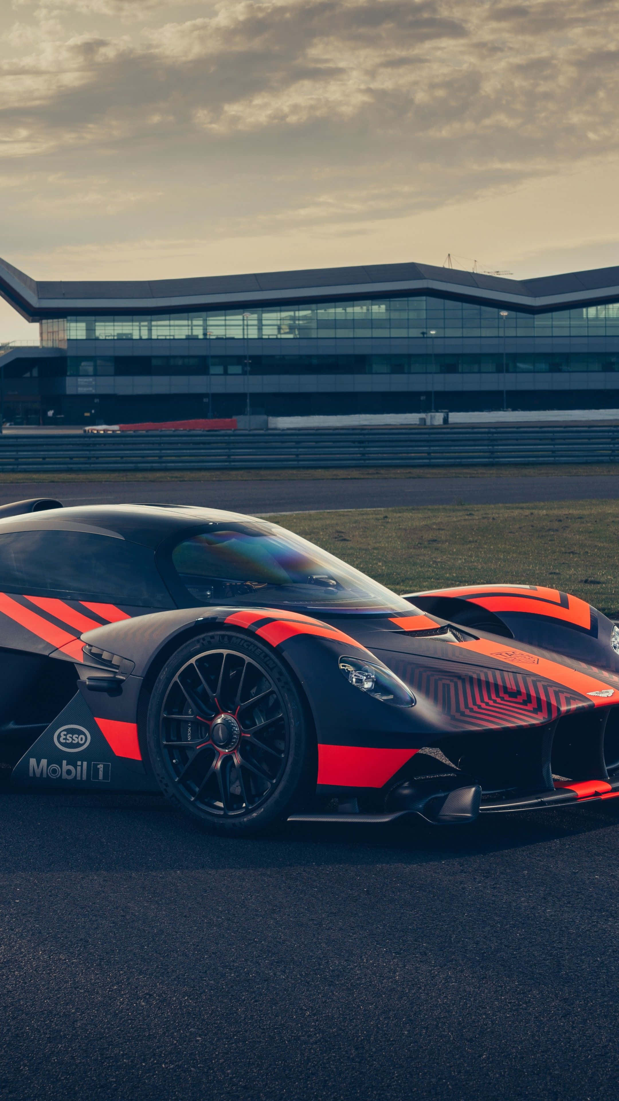 Aston Martin Valkyrie 桌布 壁紙 — 一架帶有Mobil 1標誌的時尚黑橙色賽車，在黃昏時停泊在賽道上，背景是一座現代建築。