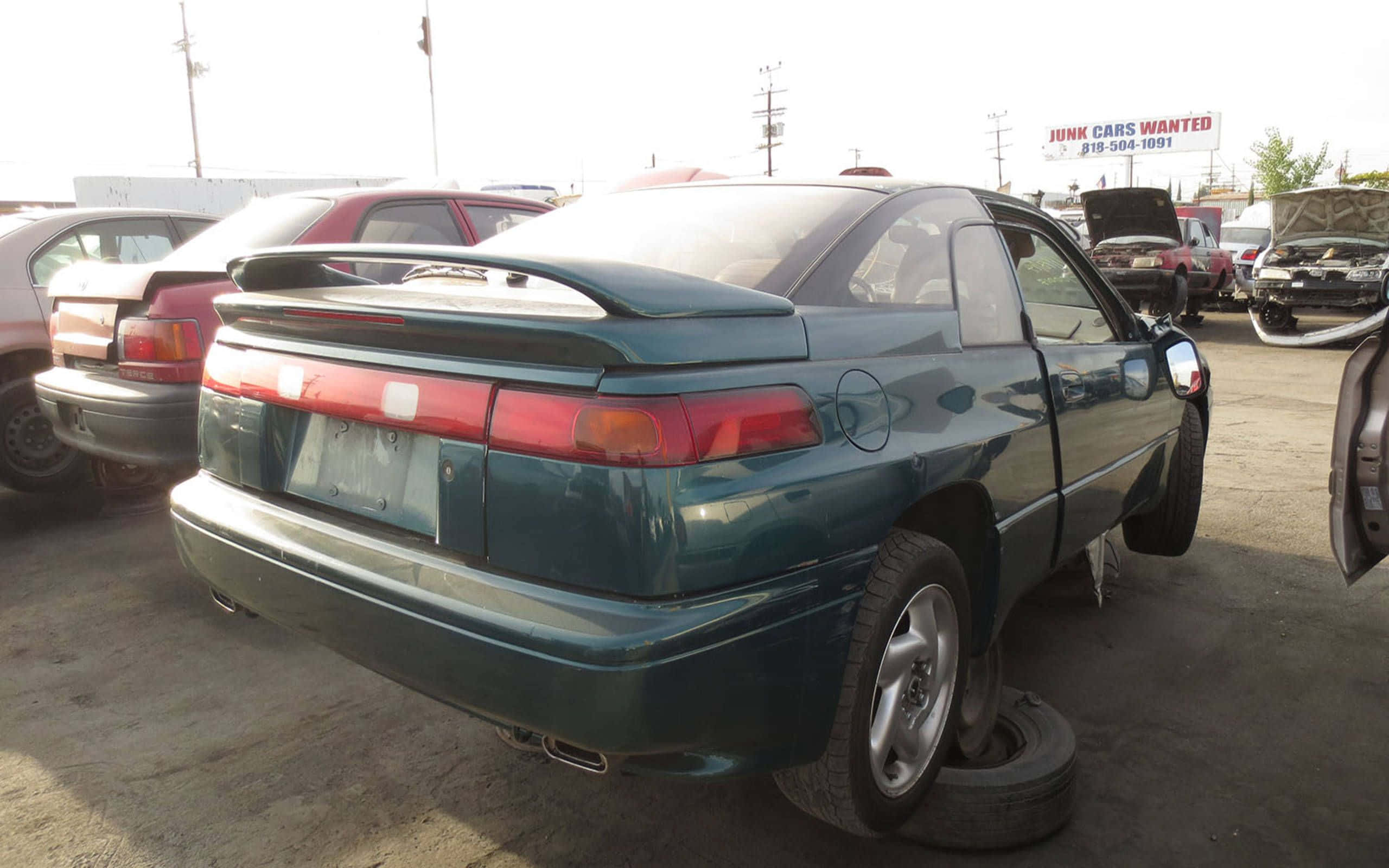 Subaru Svx 桌布 壁紙 — 一架帶有後擾流板的青綠色運動型轎跑車被遺棄在廢車場，周圍是其他損毀的汽車，在明亮的日光下。