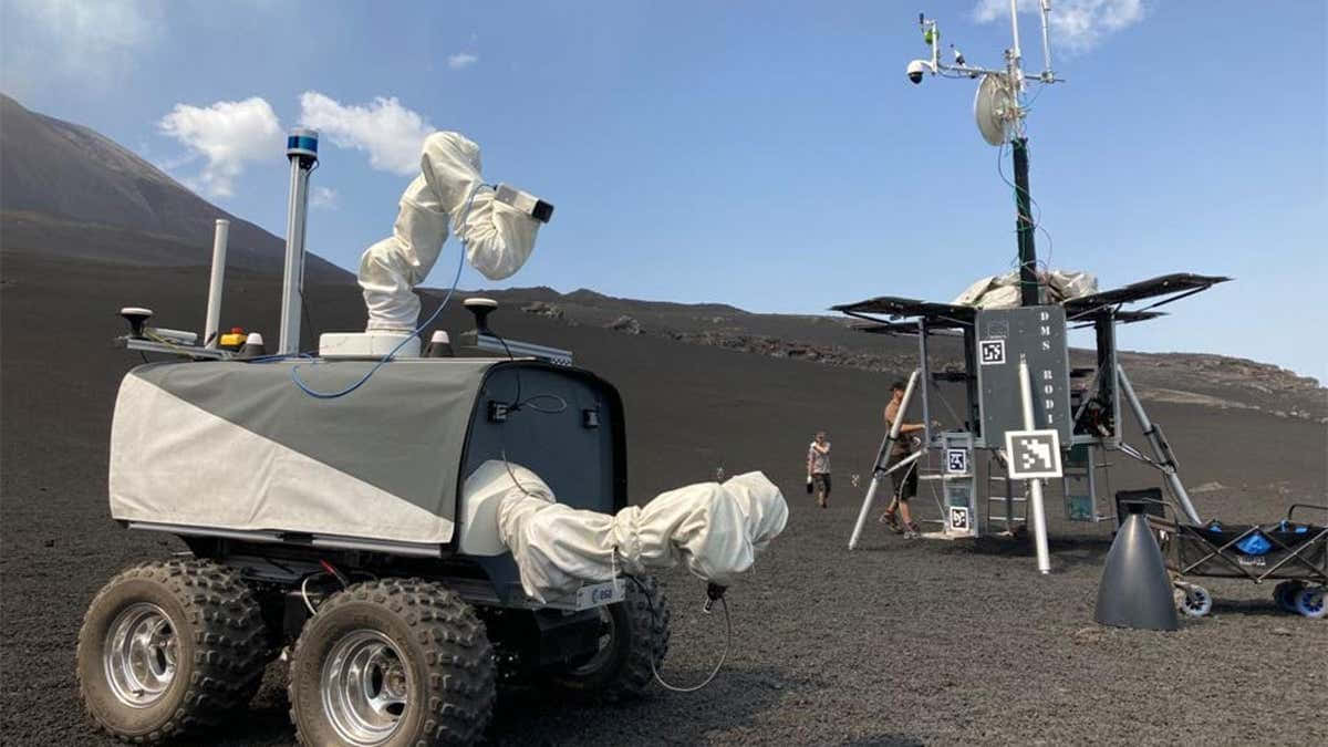 Lunar Rover 桌布 壁紙 — 一架帶有白色關節臂的機械探測車，在晴朗藍天和遠山背景下，站立在黑色火山岩場上。