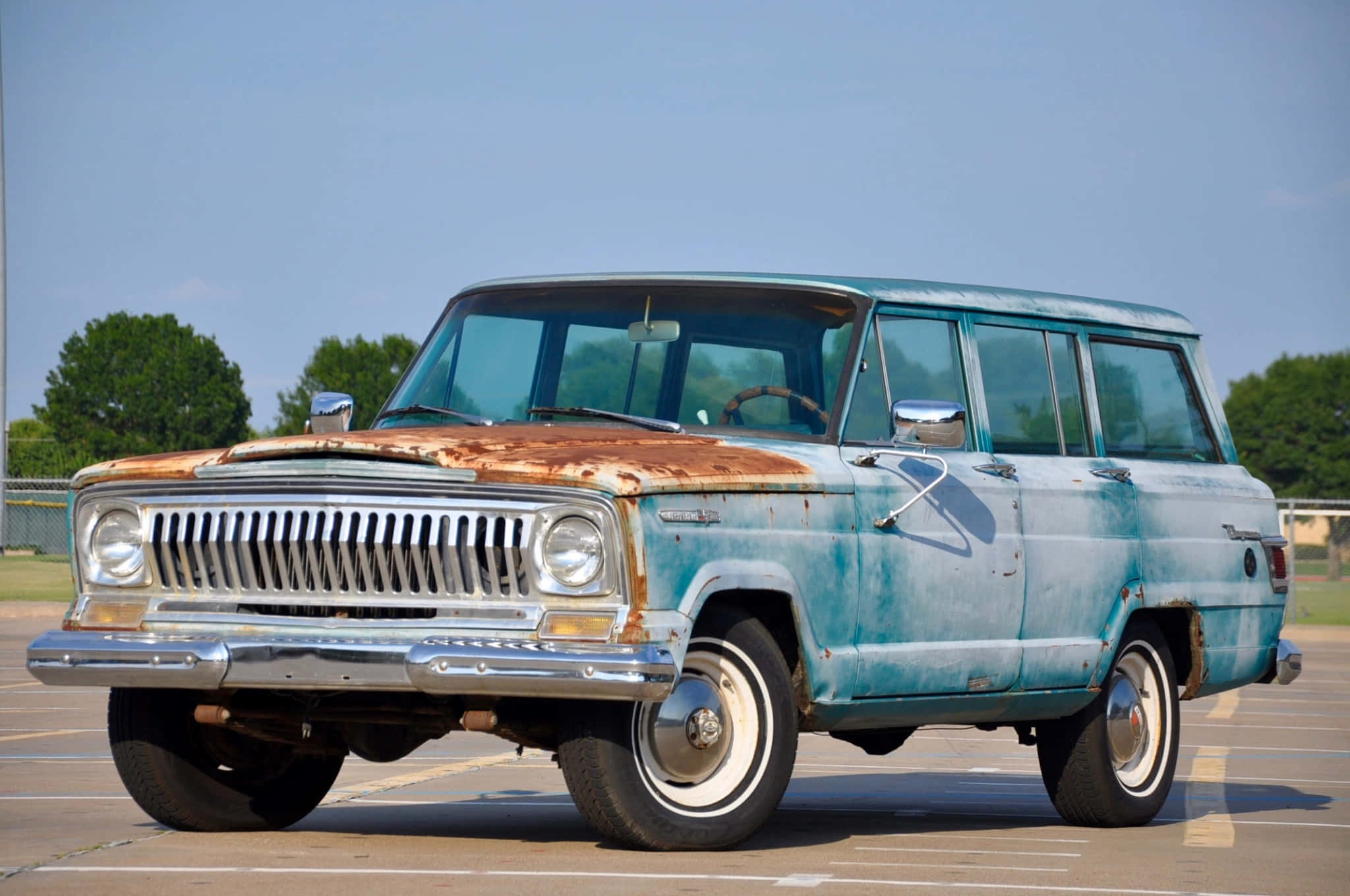 Jeep Wagoneer 桌布 壁紙 — 一架引擎蓋生鏽的復古藍色旅行車，停在陽光明媚、綠樹環繞的空地上。