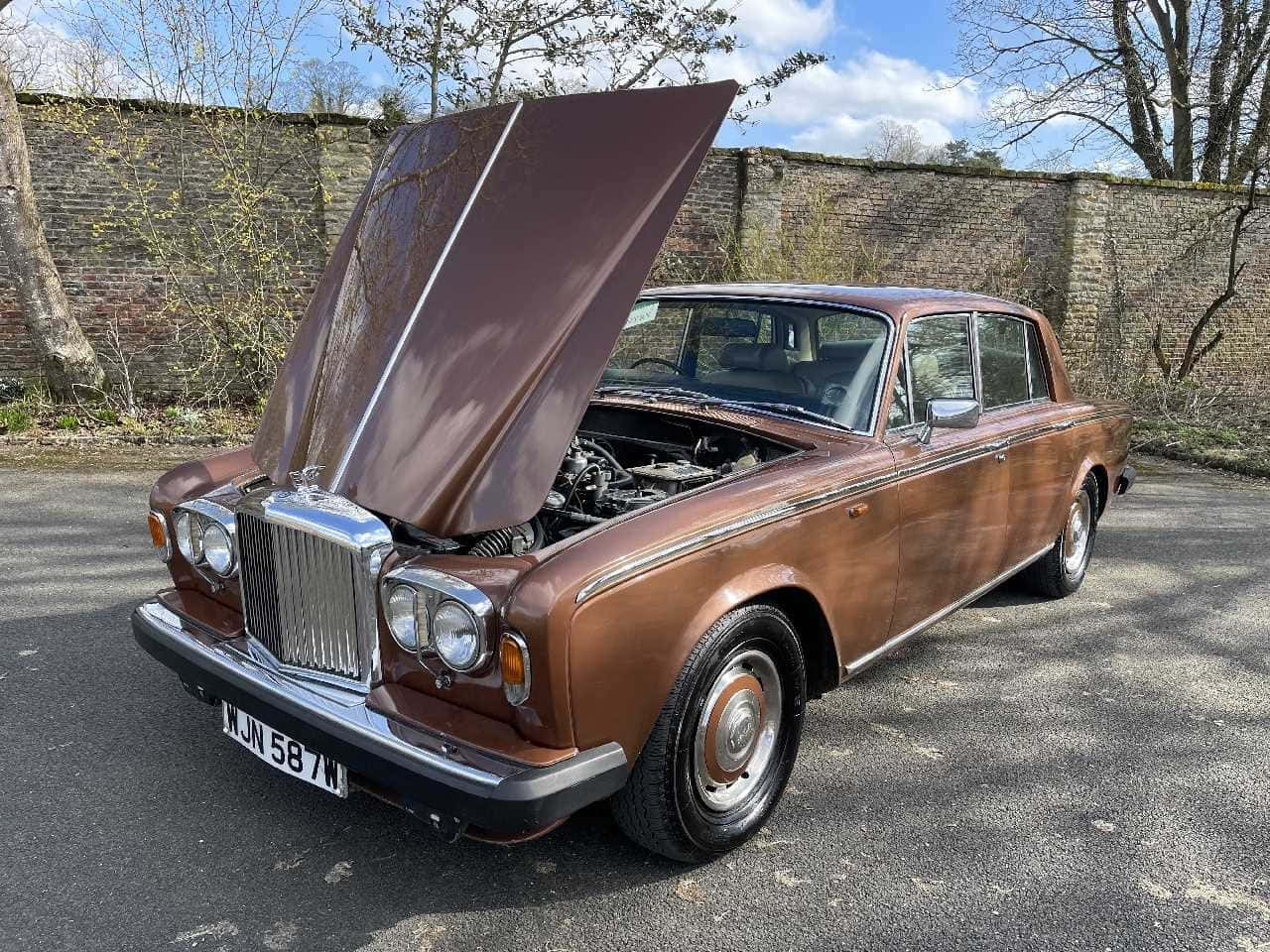 Bentley T2 桌布 壁紙 — 一架復古棕色勞斯萊斯，引擎蓋打開，停在瀝青路上，旁邊是磚牆，天空局部多雲。