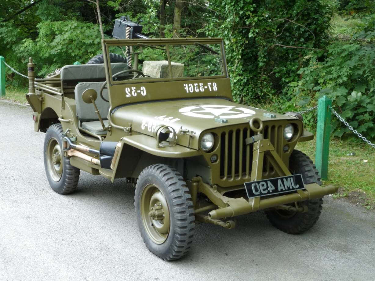 Jeep Willys 桌布 壁紙 — 一架復古橄欖綠軍用吉普車，裝有機槍，停泊在碎石路上，周圍環繞著茂密的綠色植物。