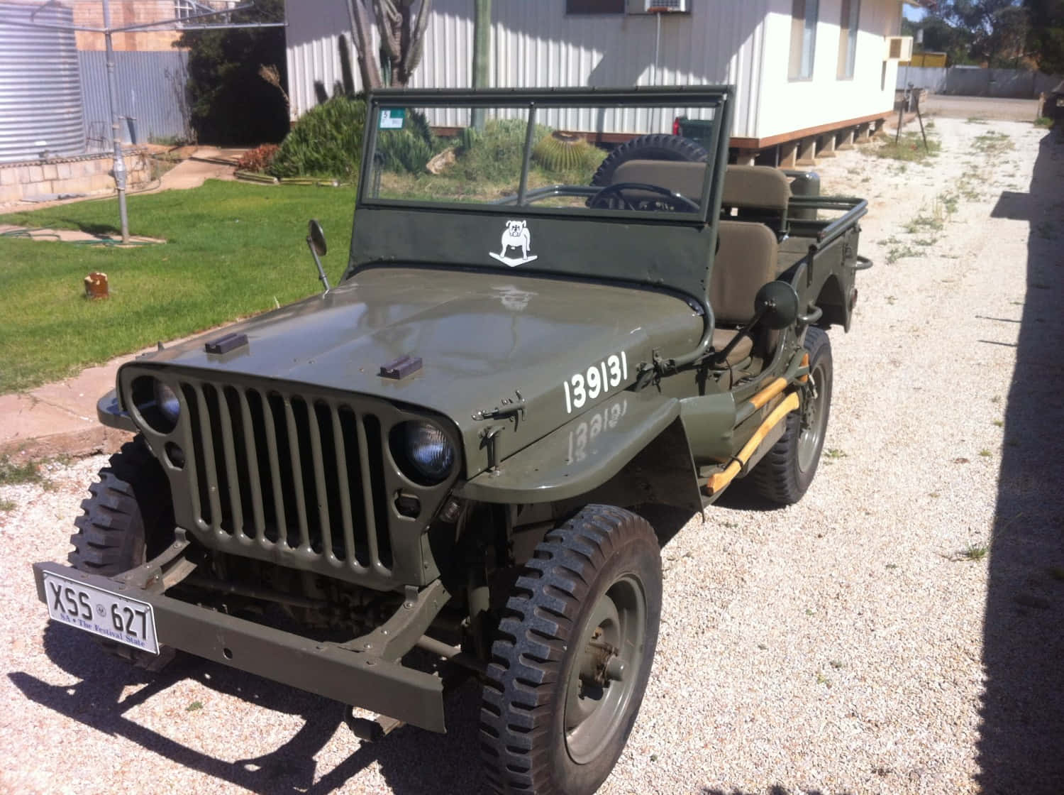 Jeep Willys 桌布 壁紙 — 一架復古橄欖綠軍用吉普車停泊在白色房屋前的碎石路上，周圍有草地和樹木，陽光普照。