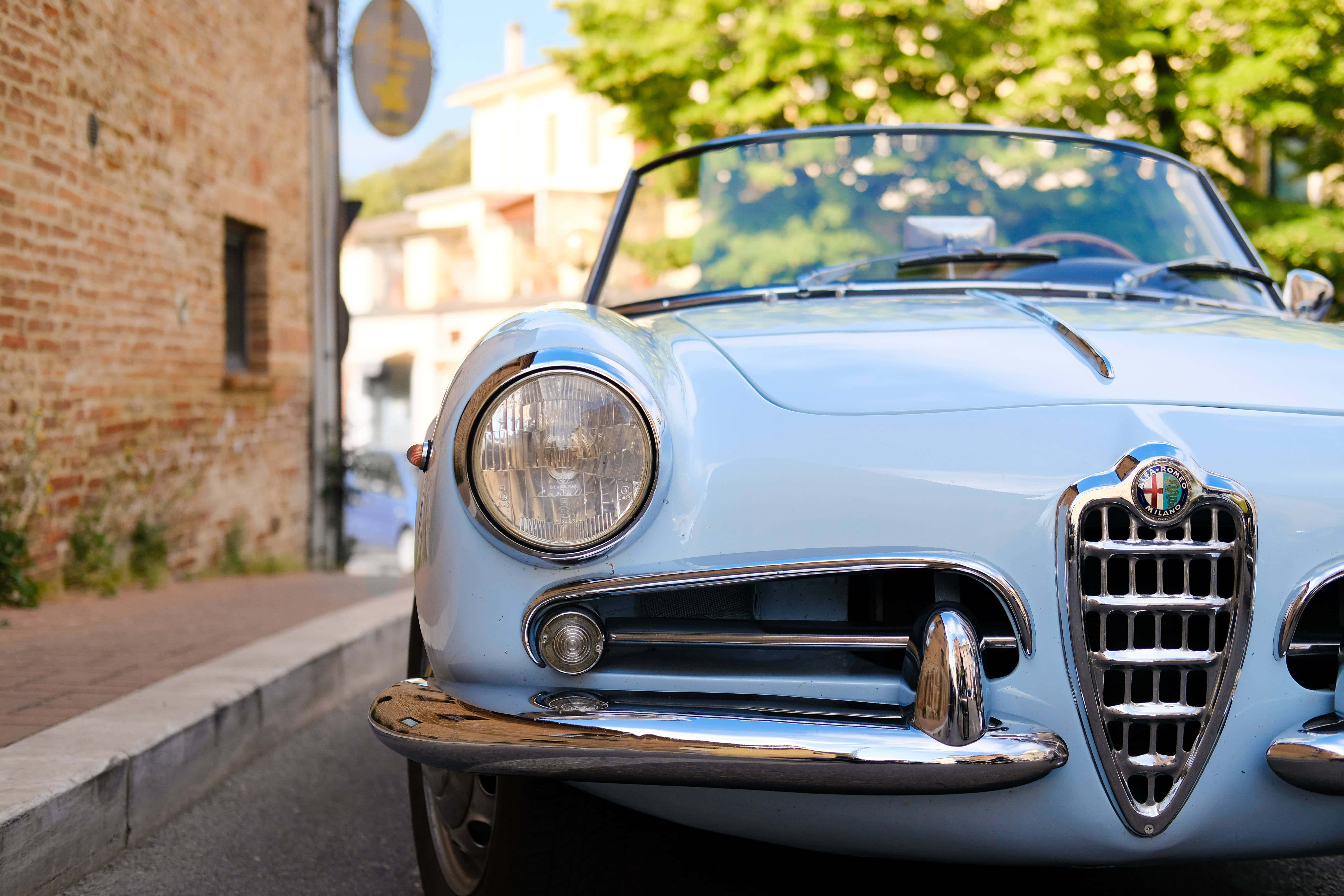 Alfa Romeo Spider 桌布 壁紙 — 一架復古淺藍色愛快羅密歐蜘蛛跑車停泊在陽光普照的鵝卵石街道上，旁邊是磚牆和綠樹。