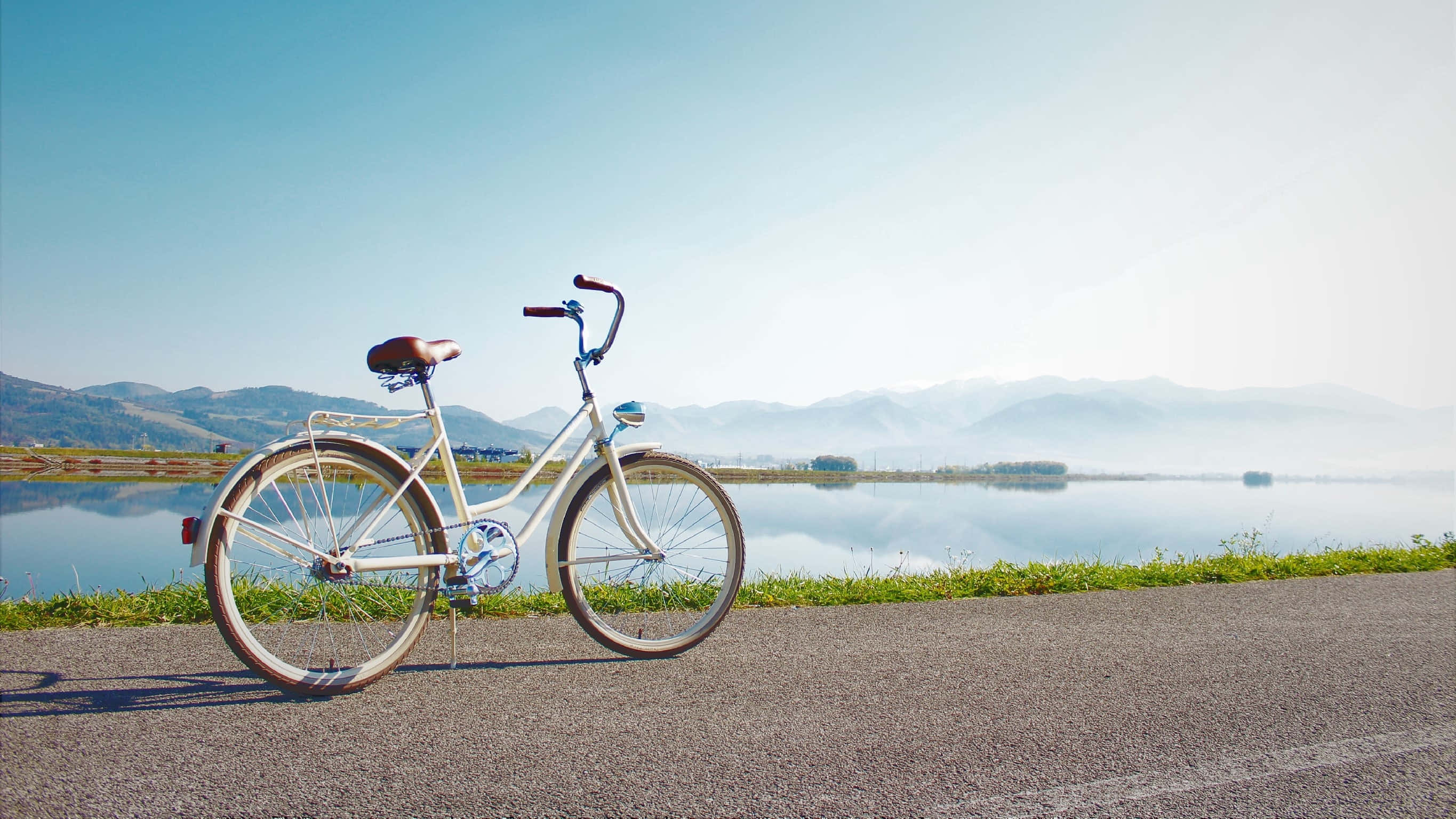 Bicycle 桌布 壁紙 — 一架復古白色單車停泊在平靜湖泊旁的鋪砌道路上，背景是霧氣繚繞的山脈和清澈的藍天。