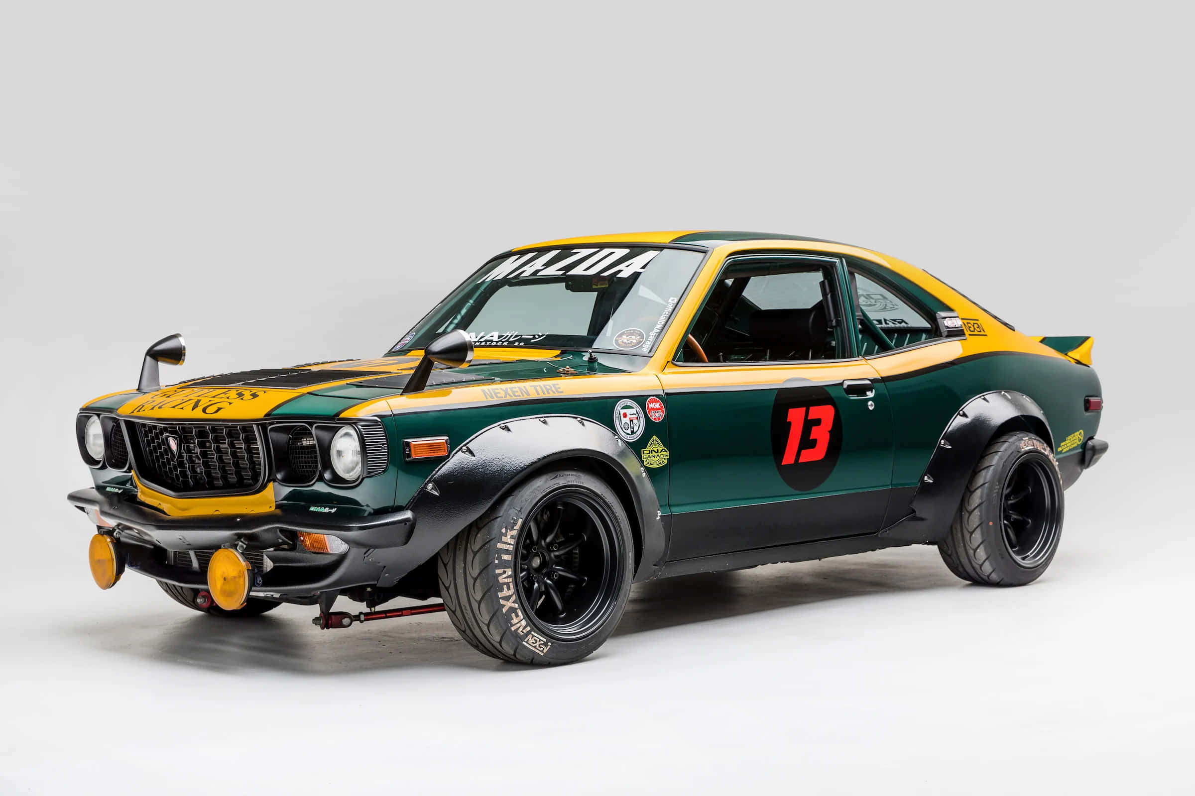 Mazda Rx-3 桌布 壁紙 — 一架復古綠色和黃色的萬事得RX-3拉力賽車，帶有13號和賽車貼紙，擺放在純白色背景前。