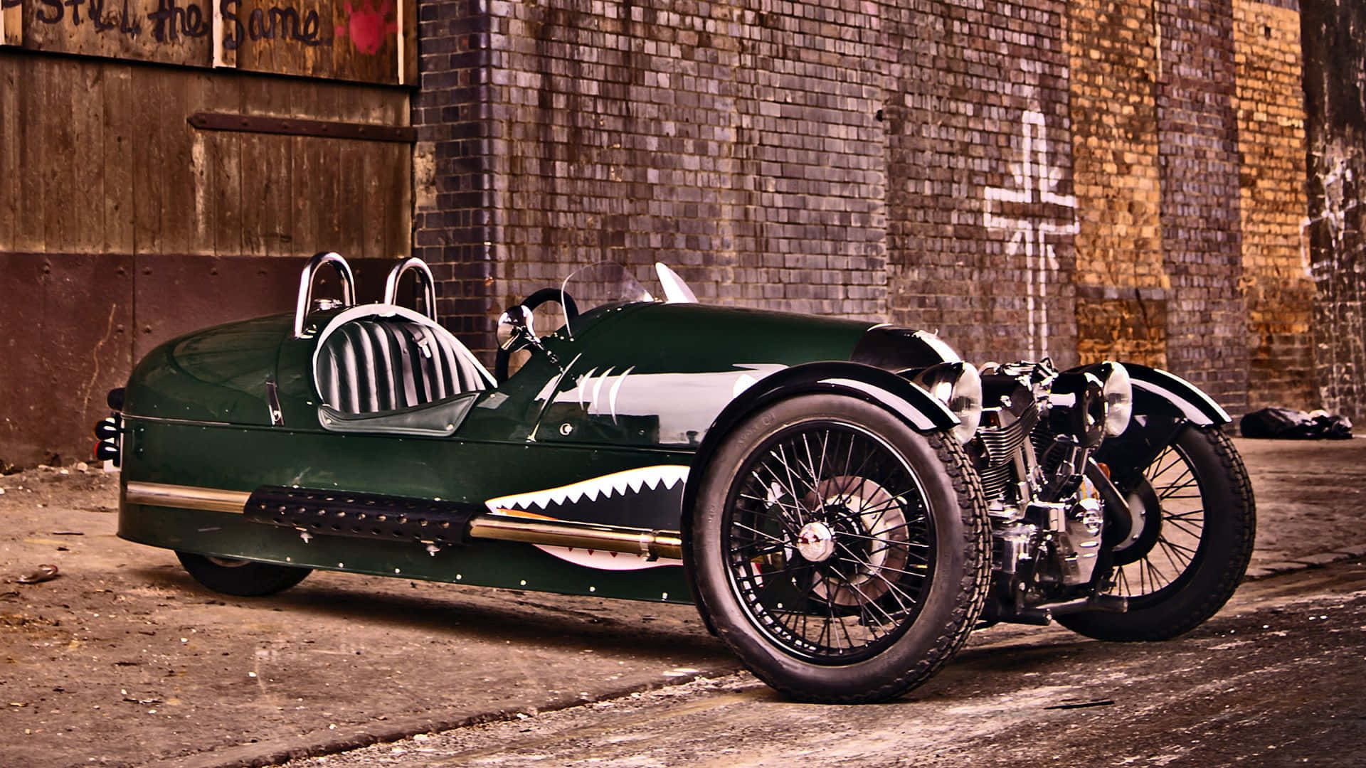 Morgan 桌布 壁紙 — 一架復古綠色賽車，帶有鯊魚嘴格柵，停泊在佈滿塗鴉的風化磚牆旁。