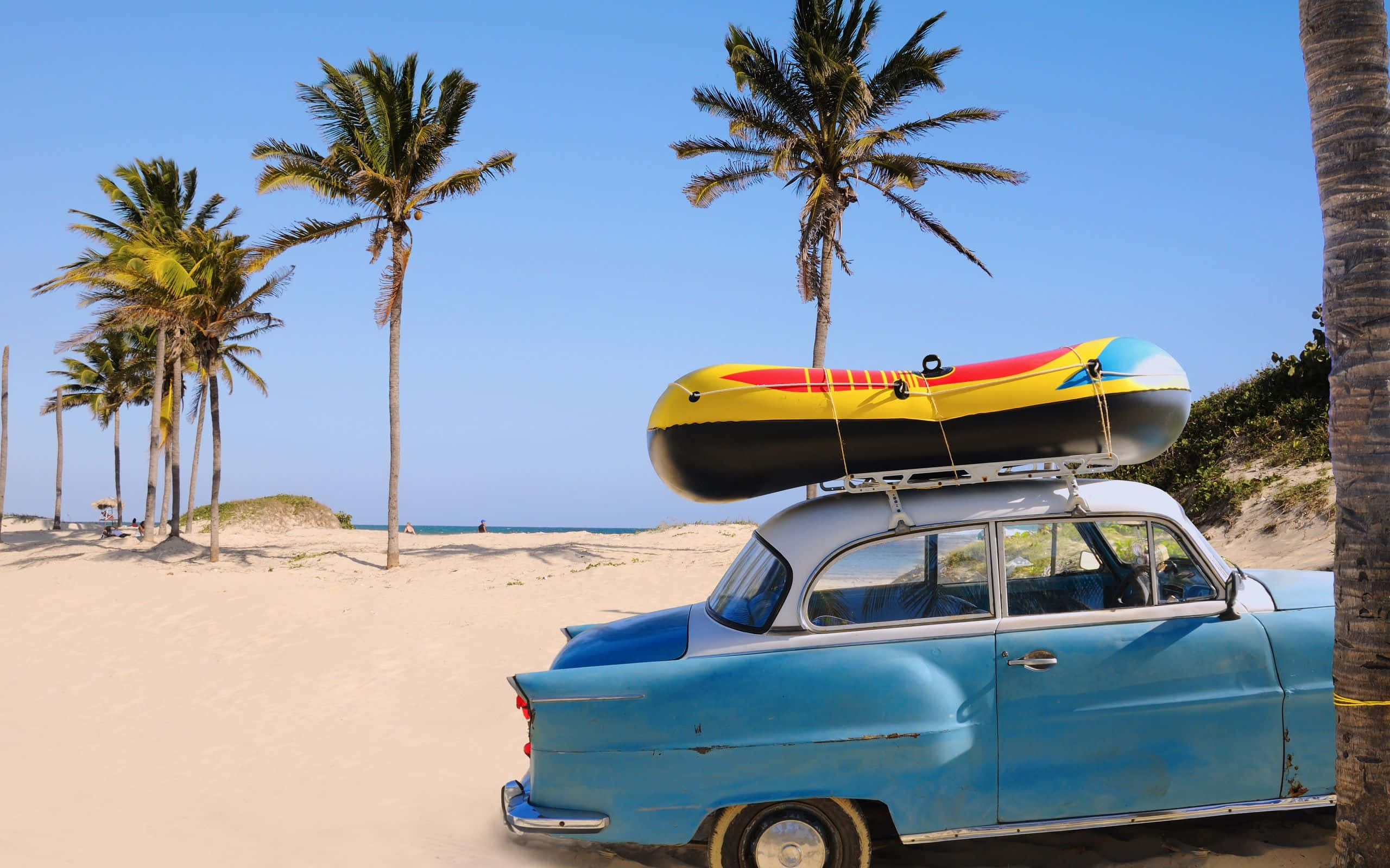 Beach Camping 桌布 壁紙 — 一架復古藍色汽車，車頂綁著色彩繽紛的充氣筏，停泊在沙灘上，周圍是高高的棕櫚樹。