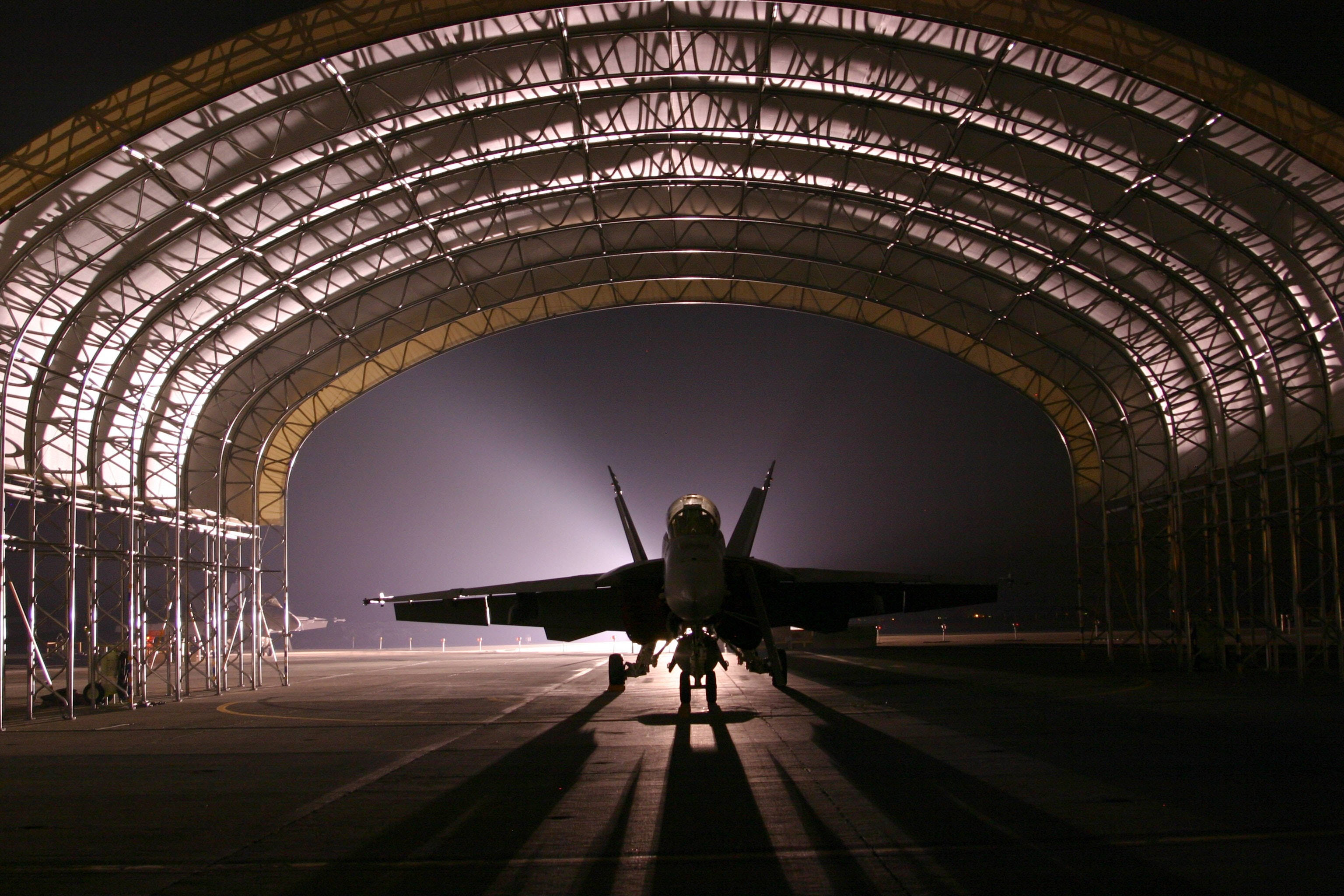 飛機 桌布 壁紙 — 一架戰鬥機在夜間於巨大機庫內呈現剪影，上方燈光投射出長長的陰影。