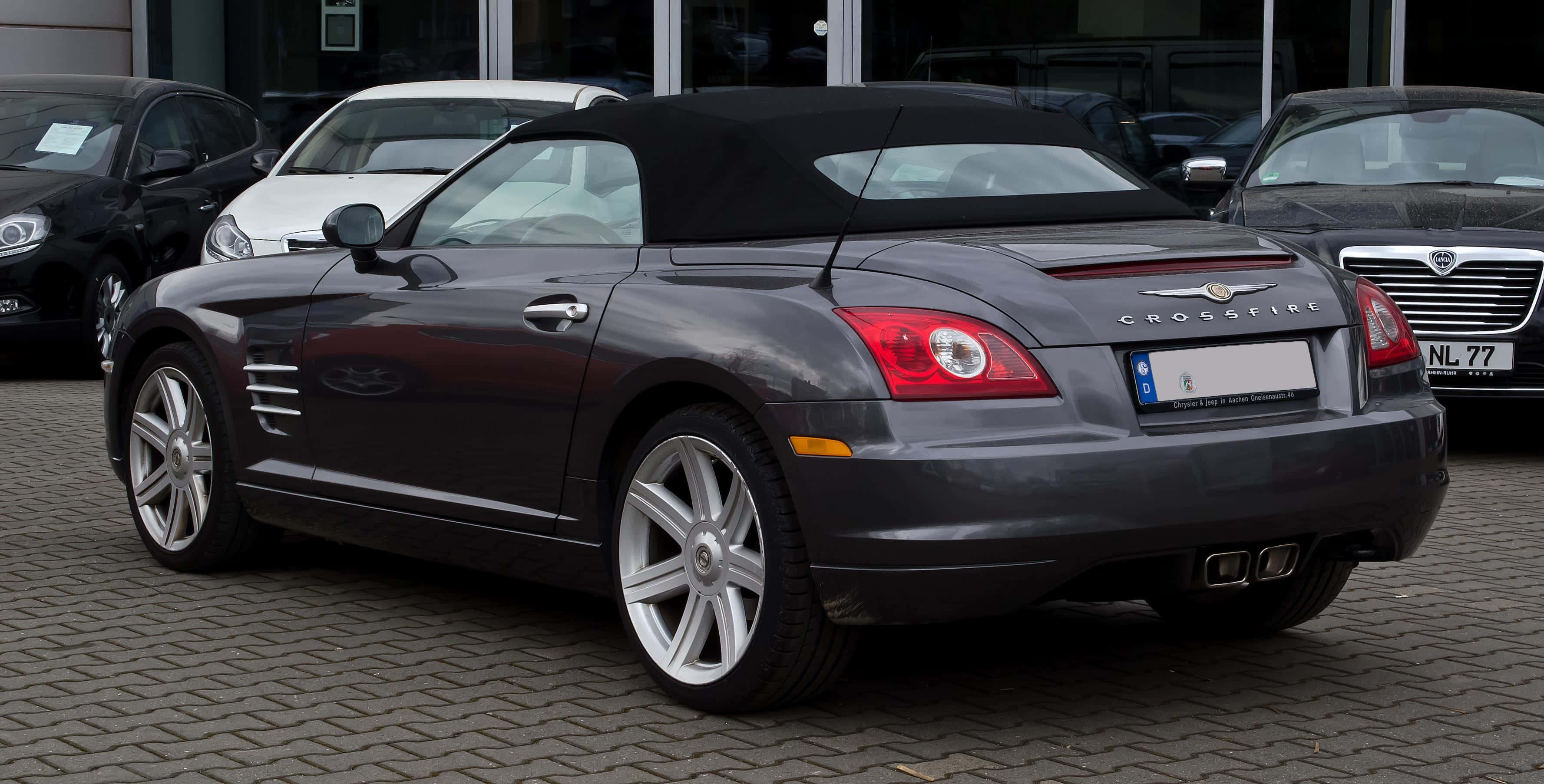 Chrysler Crossfire 桌布 壁紙 — 一架時尚嘅深灰色 Chrysler Crossfire 開篷車停泊喺鵝卵石停車場，背景有其他車輛。