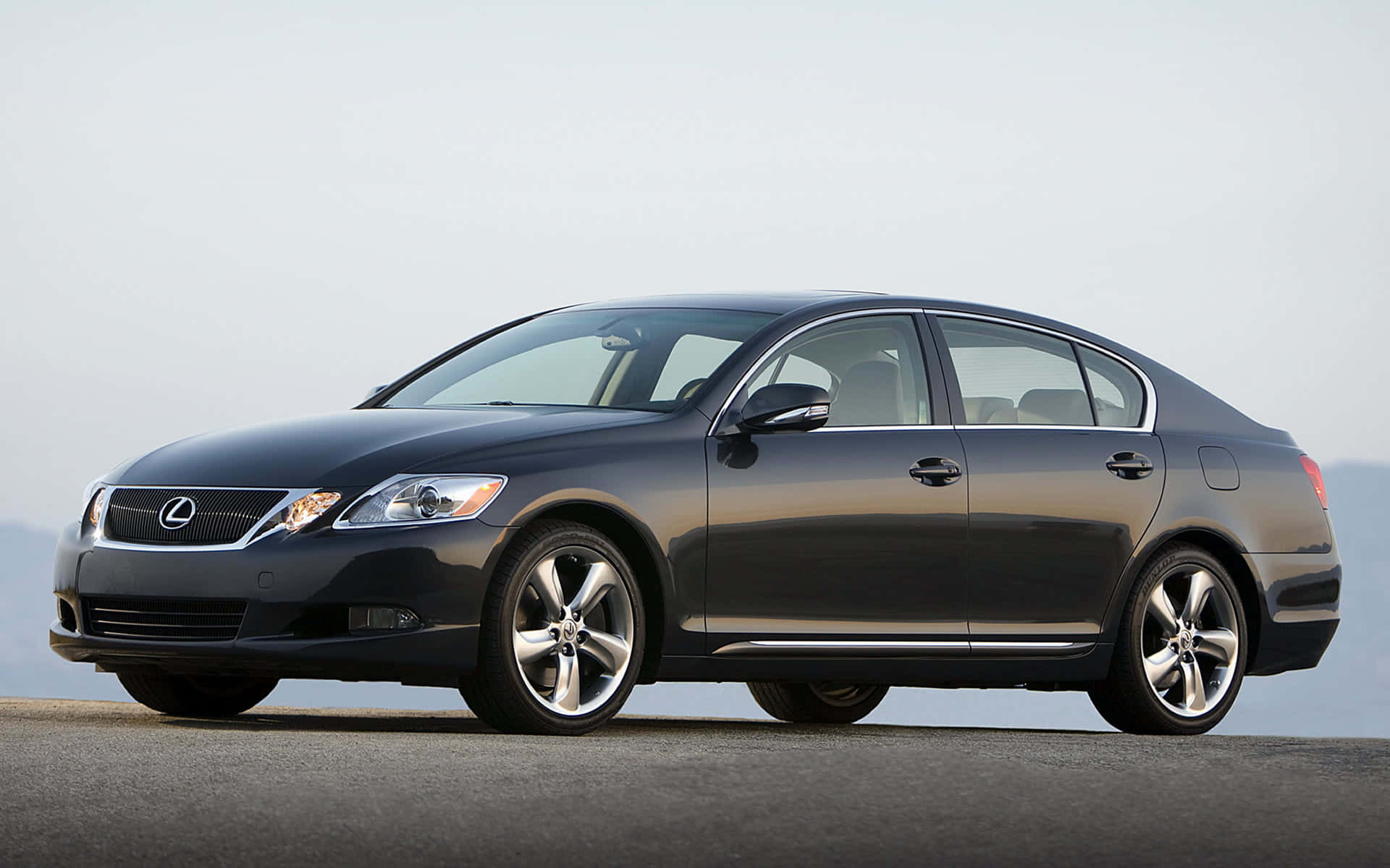 Lexus Gs 桌布 壁紙 — 一架時尚嘅深灰色 Lexus 房車停泊喺開放嘅鋪砌表面上，喺淡色天空下有一片朦朧嘅遠山山脈。