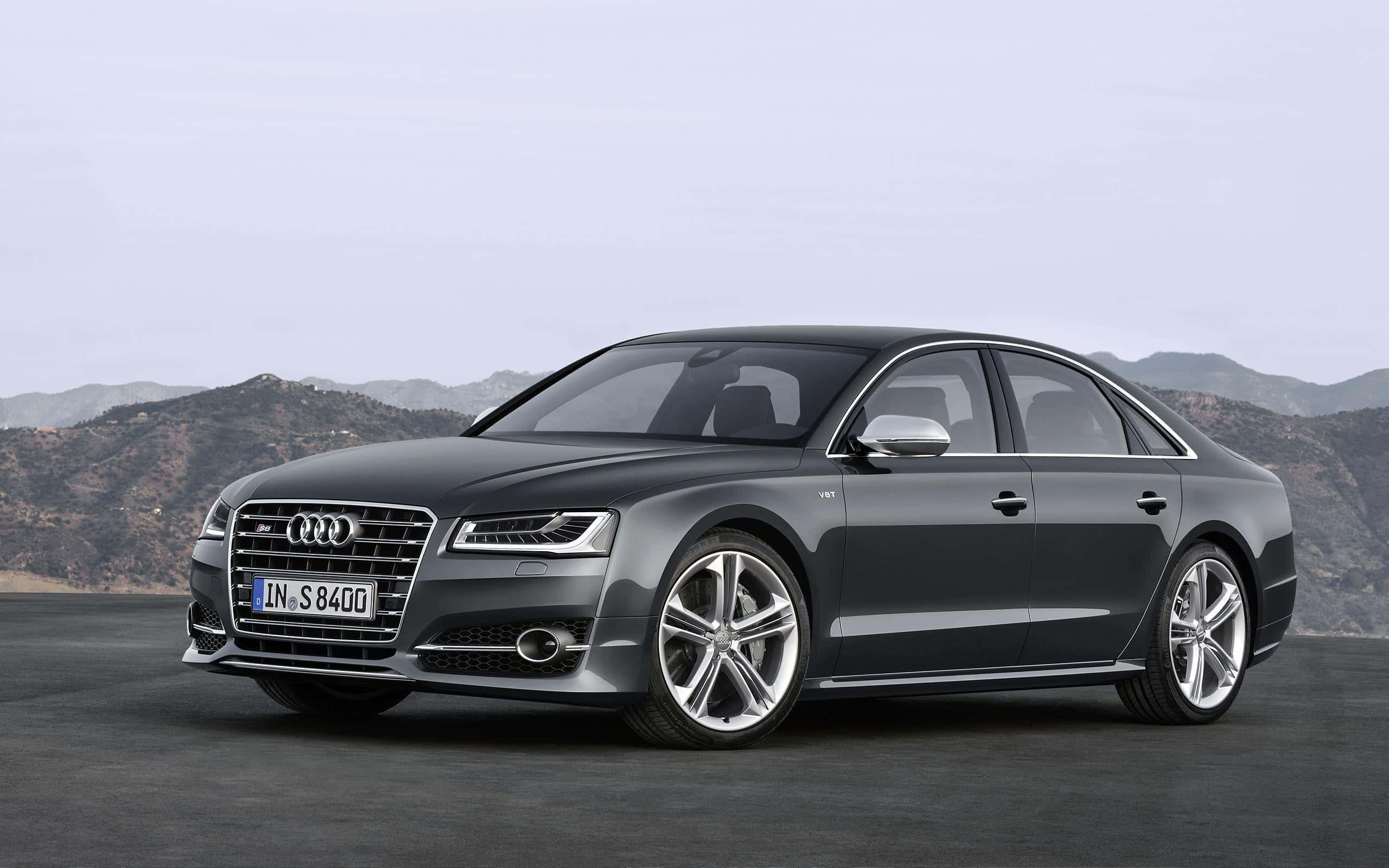 Audi S8 桌布 壁紙 — 一架時尚嘅深灰色奧迪S8房車停泊喺鋪砌嘅路面上，遠處有山脈，天空蒼白。