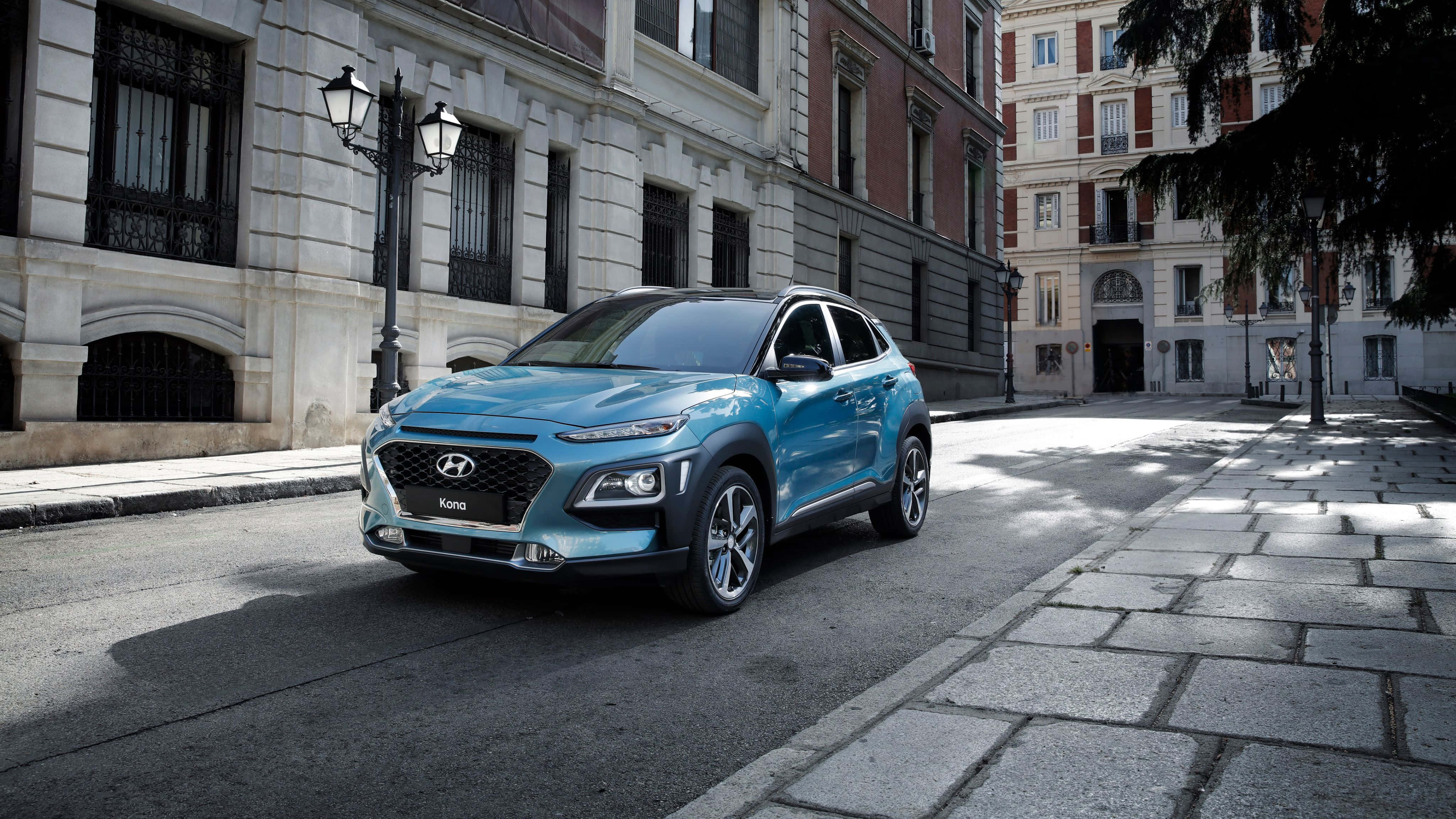Hyundai Kona 桌布 壁紙 — 一架時尚嘅青綠色Hyundai Kona SUV停泊喺陽光普照嘅鵝卵石街道上，旁邊係歷史悠久嘅歐洲建築物。