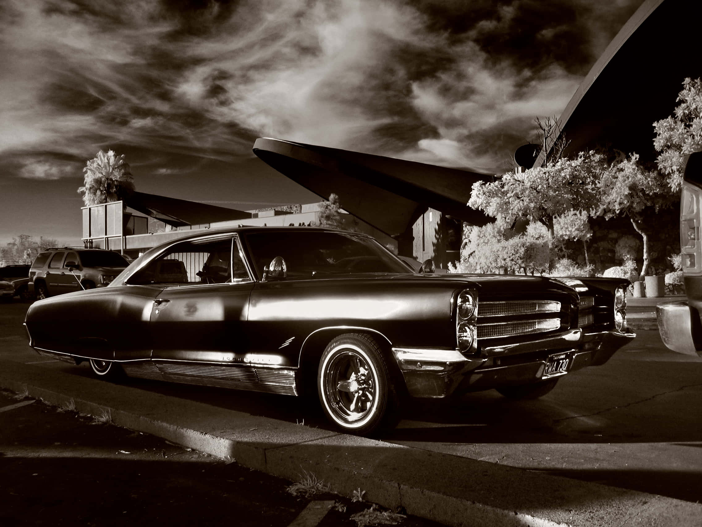 Black And White Vintage Car 桌布 壁紙 — 一架時尚復古肌肉車停泊在戲劇性的多雲天空下，旁邊是一座現代主義建築。