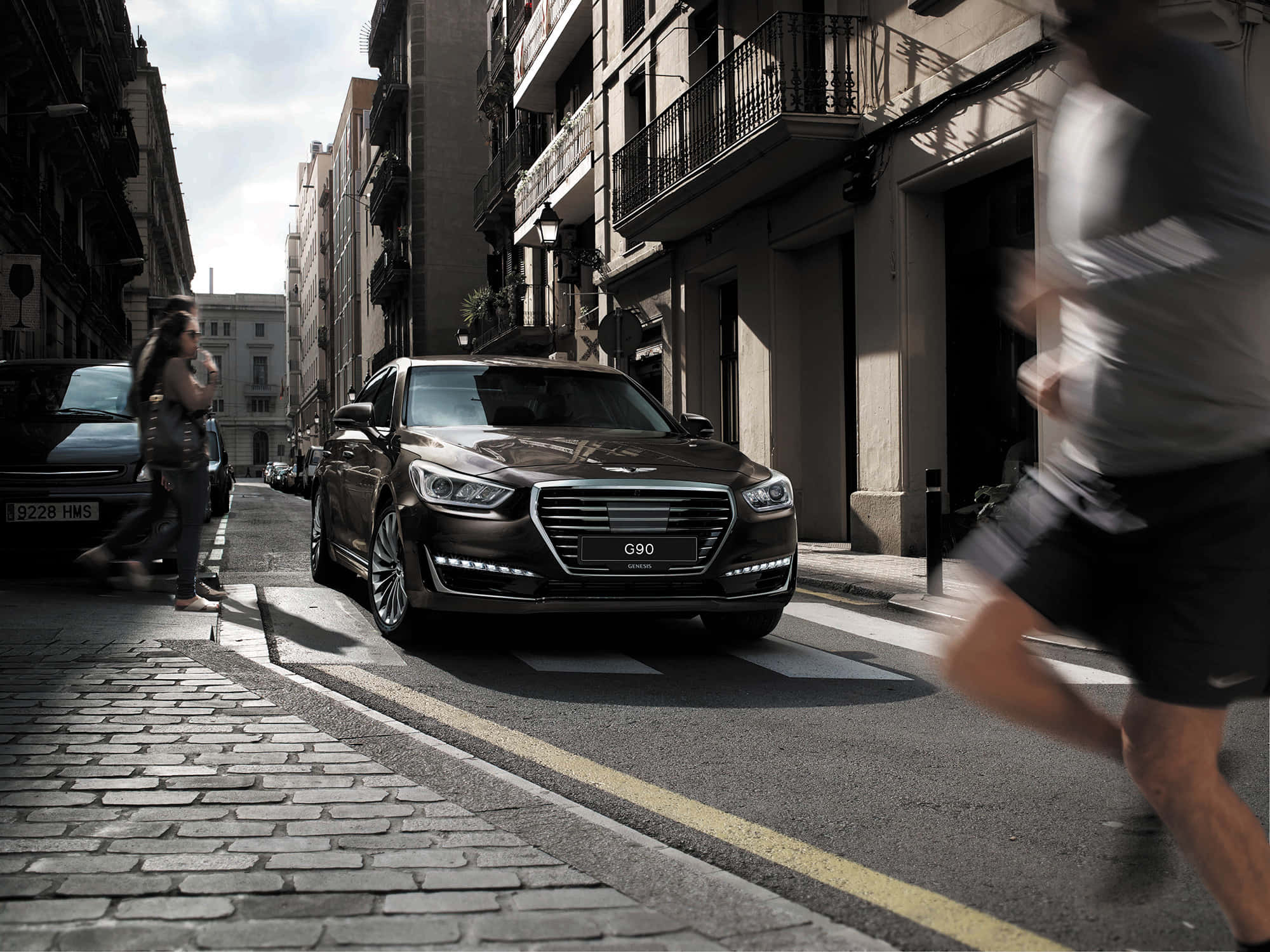 Genesis G90 桌布 壁紙 — 一架時尚深棕色Genesis G90房車停泊在陽光普照的鵝卵石街道上，模糊的行人正在走動。