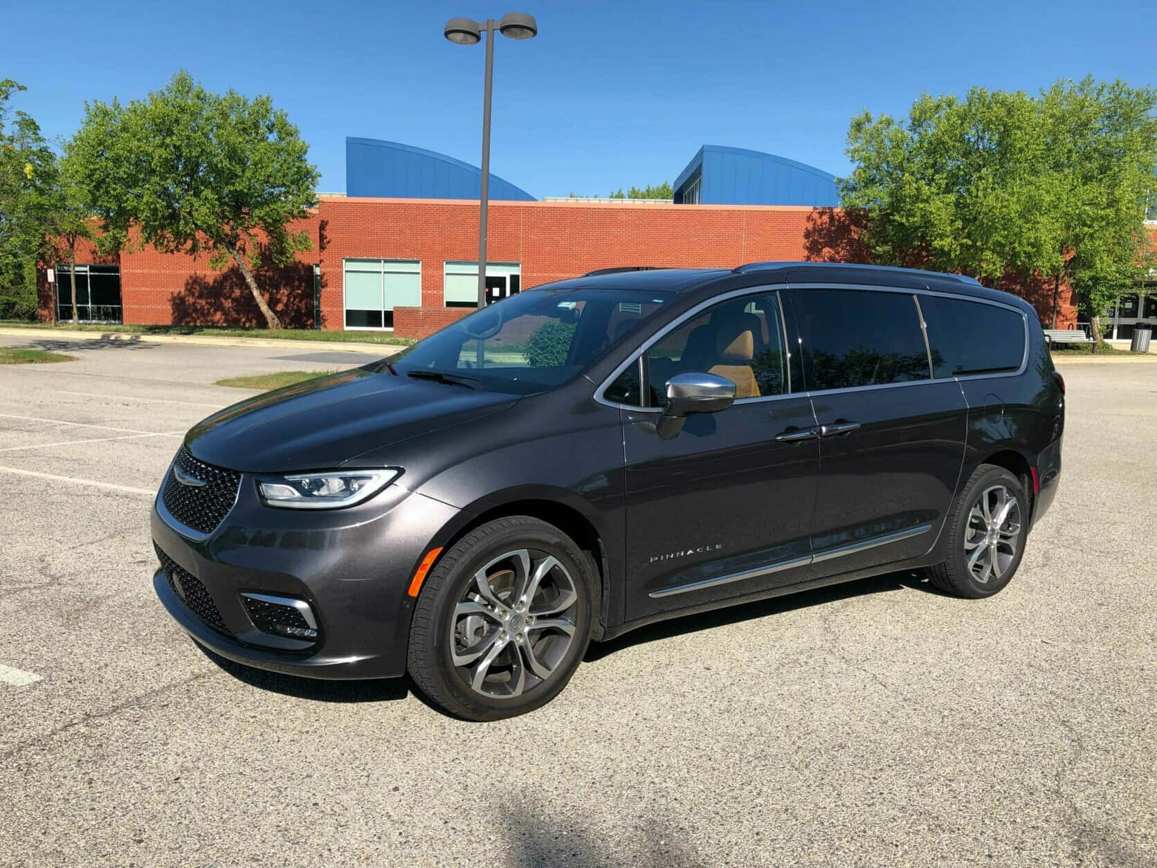Chrysler Pacifica 桌布 壁紙 — 一架時尚深灰色 Chrysler Pacifica 小型貨車停泊在陽光普照的瀝青停車場，背景有磚砌建築物和綠樹。