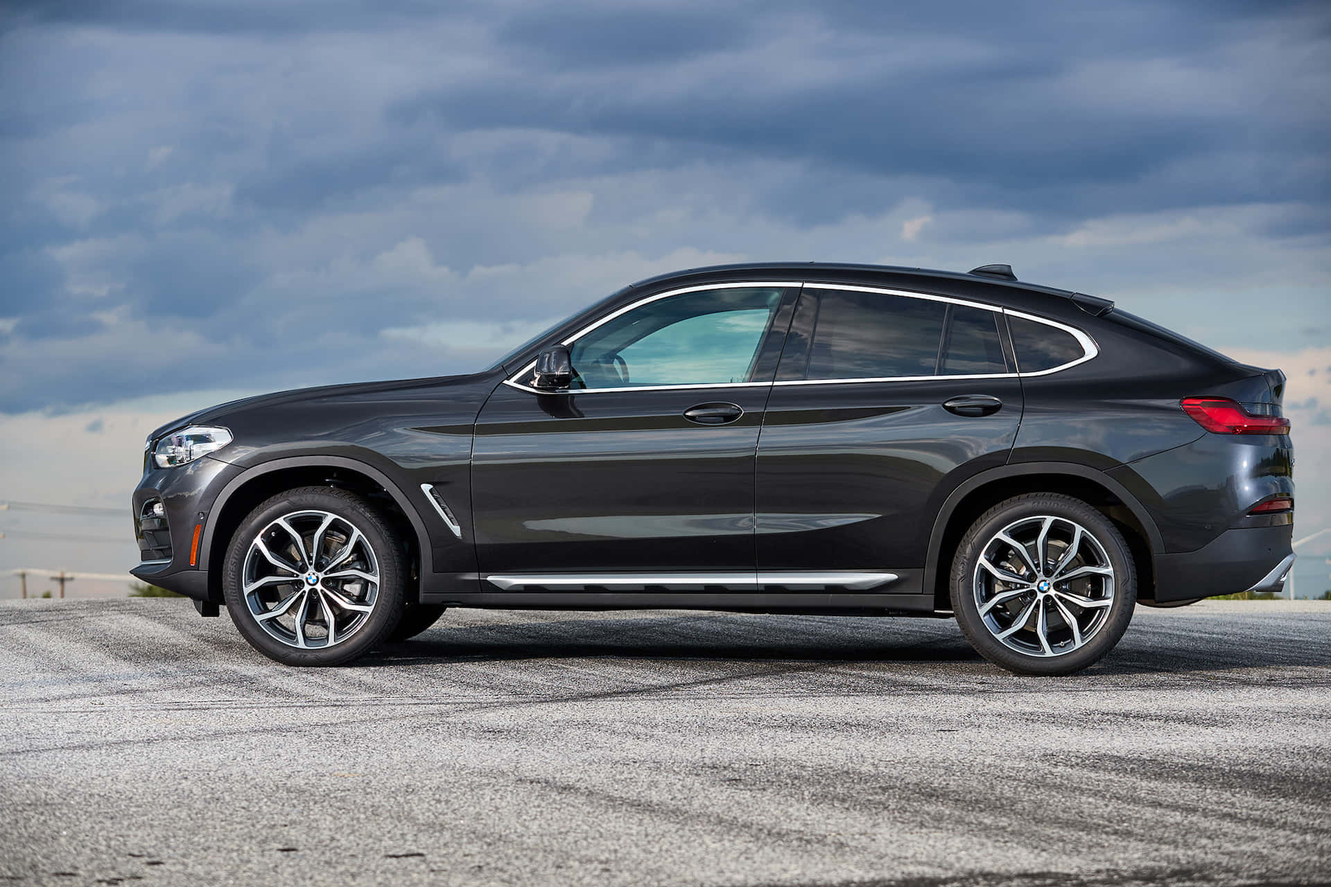 Bmw X4 桌布 壁紙 — 一架時尚深灰色BMW X4 SUV停泊在碎石路面上，背景是戲劇性的多雲天空。