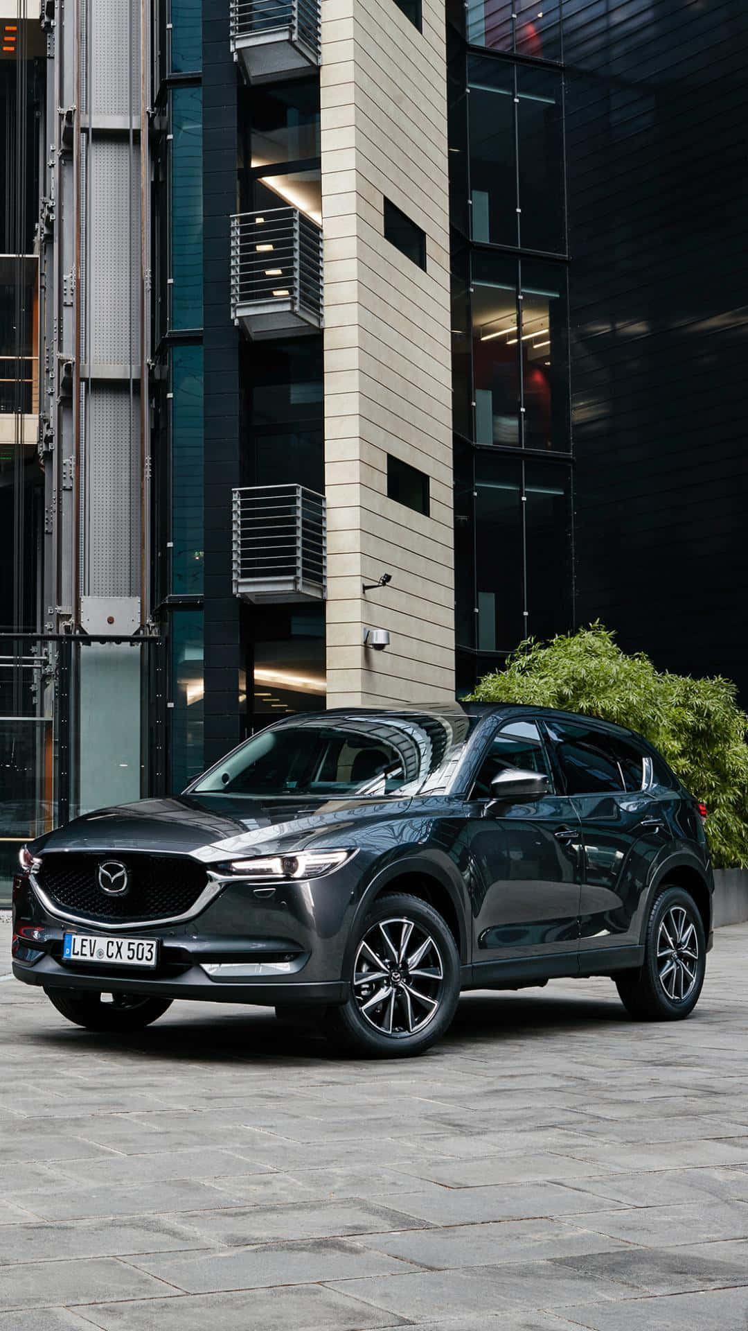 Mazda Cx-5 桌布 壁紙 — 一架時尚深灰色Mazda CX-5 SUV停泊在石頭廣場上，旁邊係一棟有綠化嘅現代玻璃同混凝土建築。