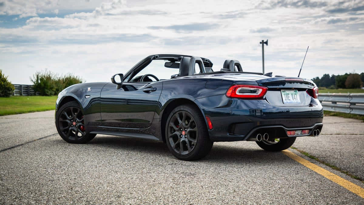 Fiat 124 Spider 桌布 壁紙 — 一架時尚深藍色開篷跑車停泊在路邊，背景是陰天和綠化。