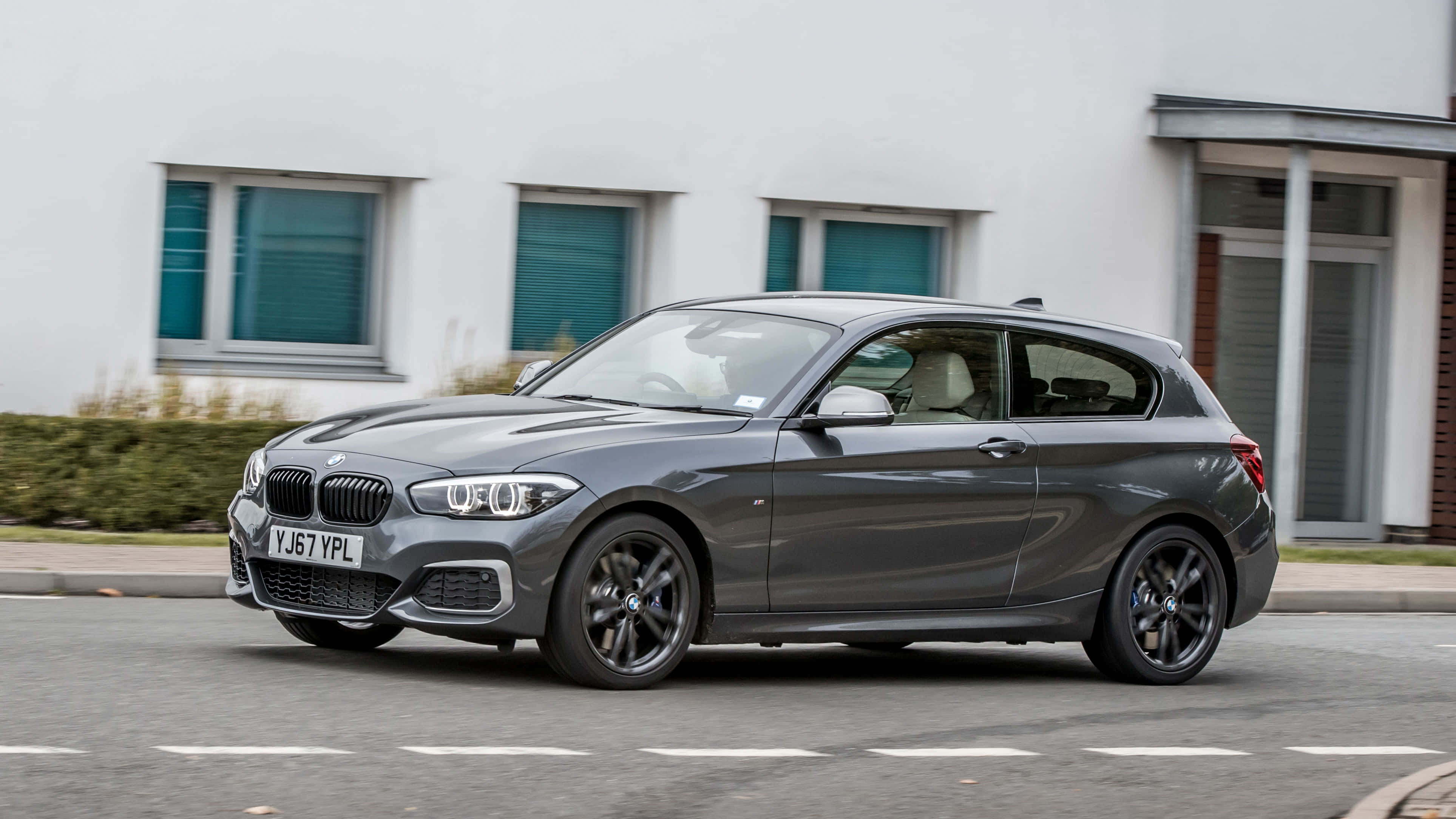 Bmw 1 Series 桌布 壁紙 — 一架時尚灰色BMW 1系掀背車駛過一棟現代白色建築，建築帶有青綠色窗戶。