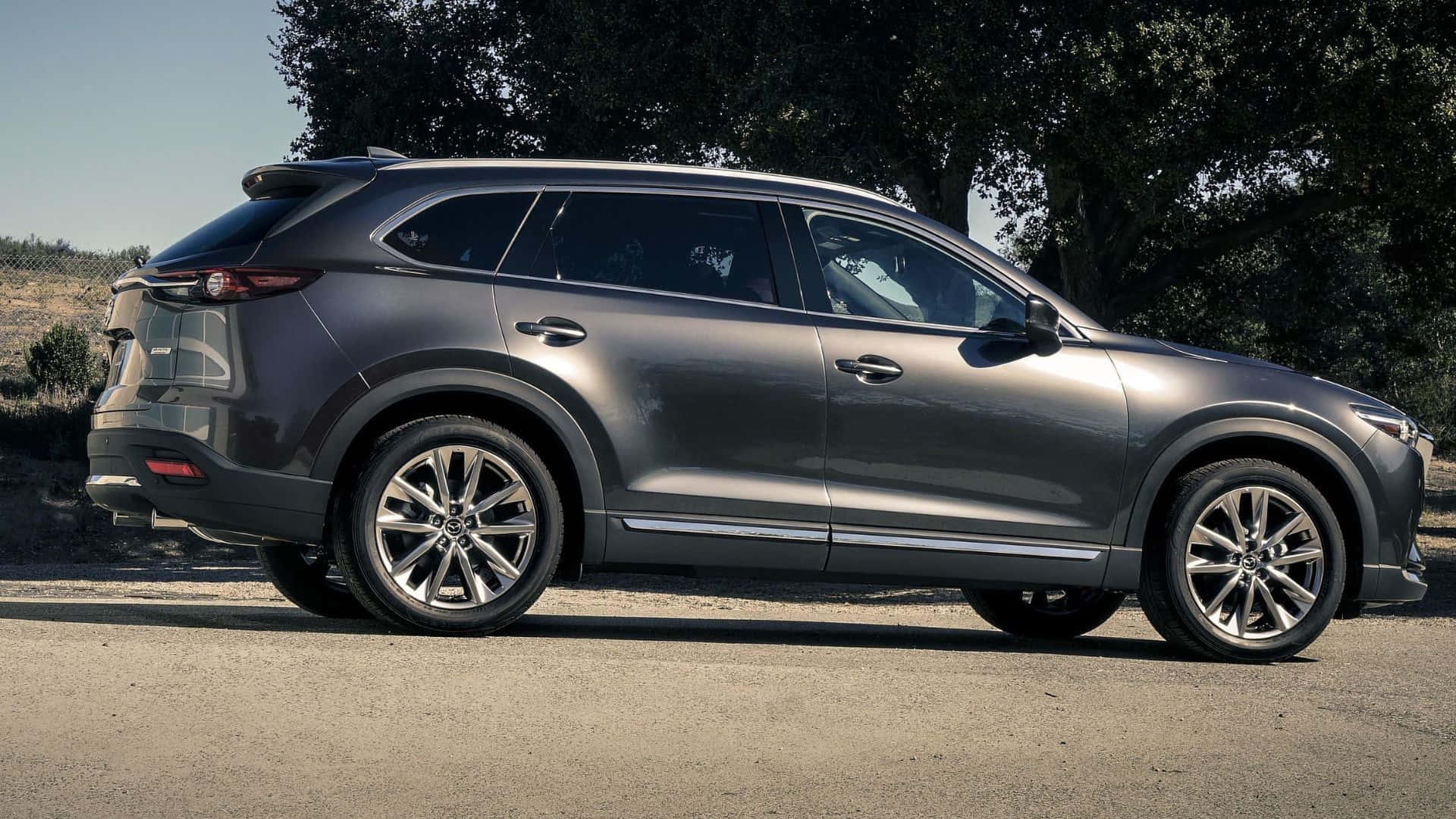 Mazda Cx-9 桌布 壁紙 — 一架時尚灰色Mazda CX-90 SUV停泊在陽光照耀的碎石路上，背景有樹木。