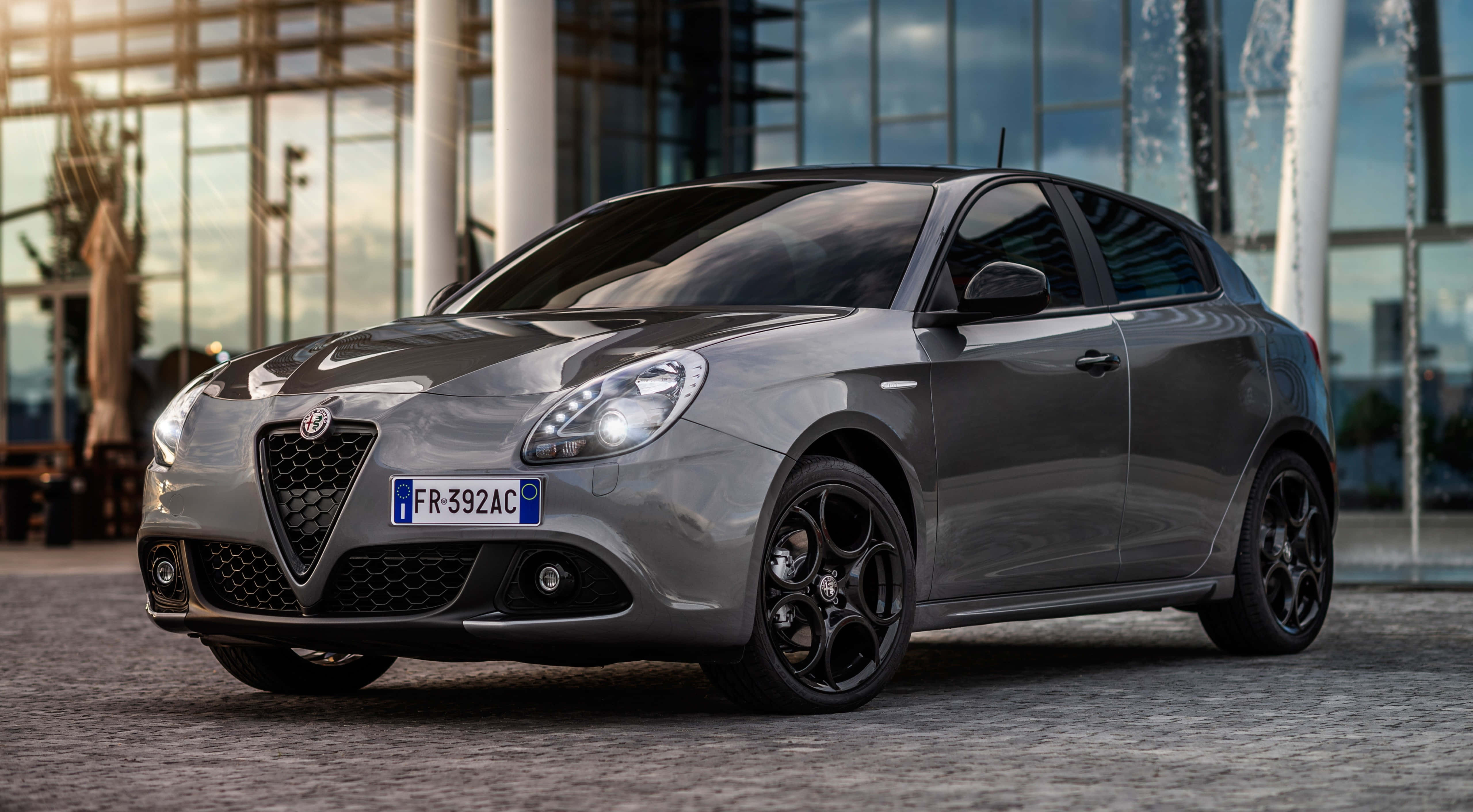 Alfa Romeo Giulietta 桌布 壁紙 — 一架時尚灰色嘅阿爾法羅密歐Giulietta停泊喺鵝卵石路上，前面係有噴泉嘅現代玻璃建築。