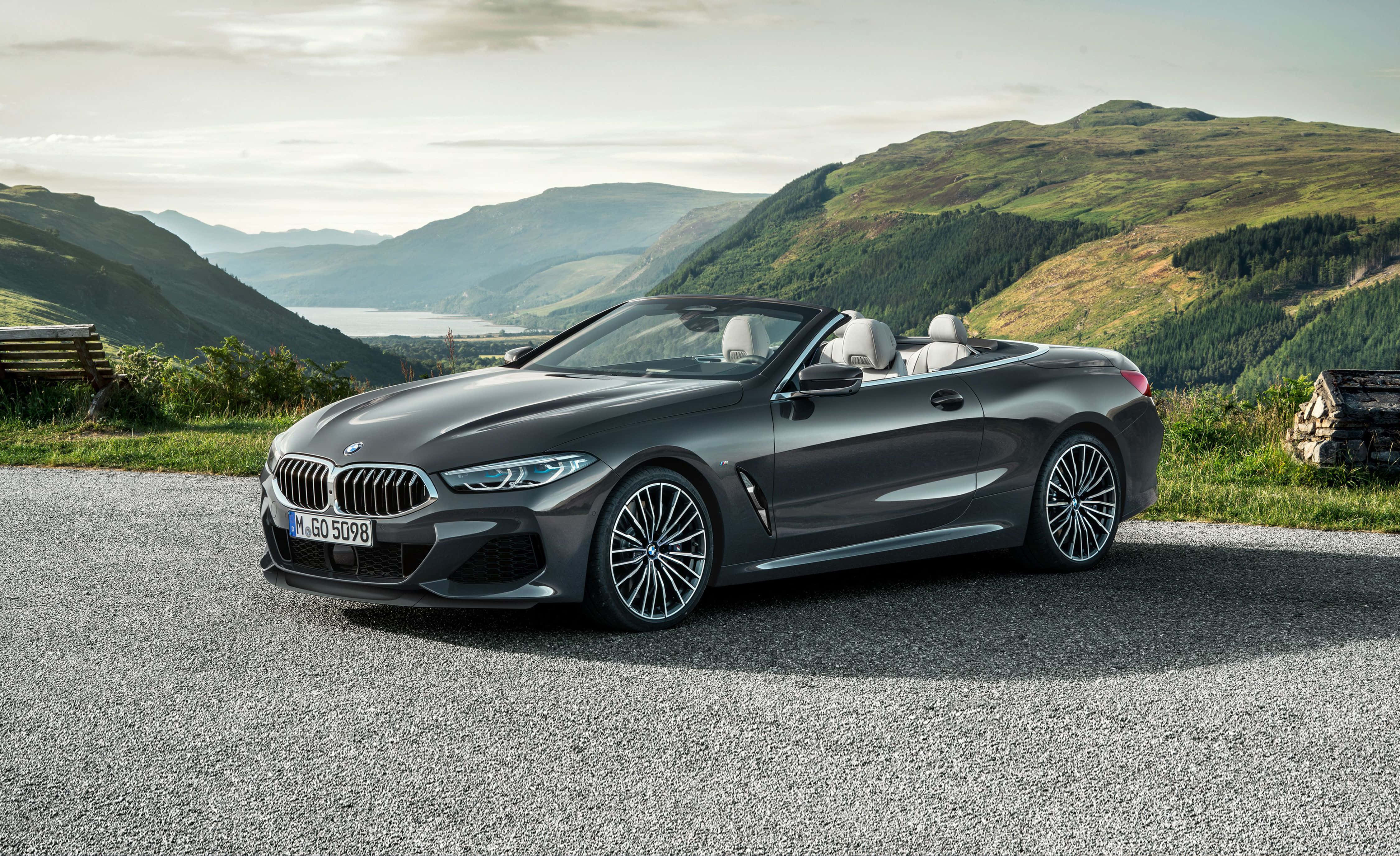 Bmw 8 Series 桌布 壁紙 — 一架時尚灰色寶馬開篷車停泊在碎石路上，周圍是起伏的綠色山丘和遠處的湖泊，天空柔和。