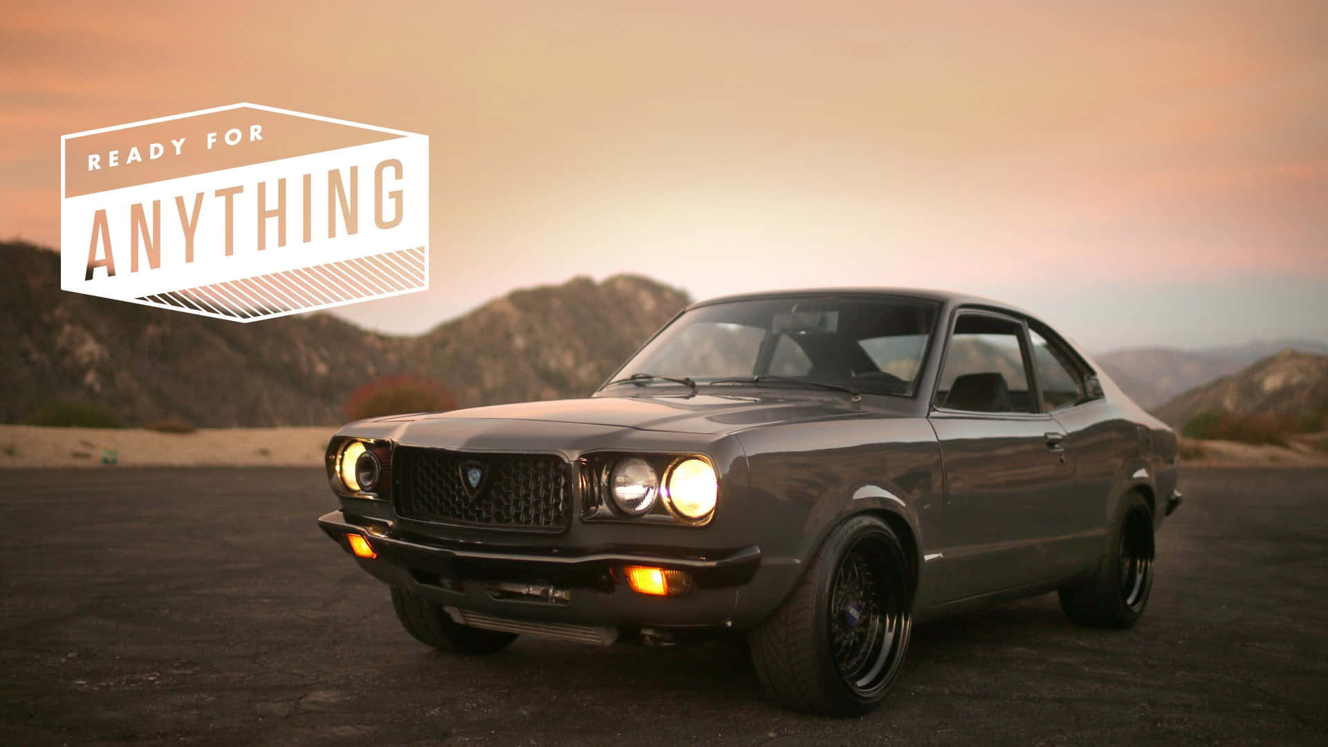Mazda Rx-3 桌布 壁紙 — 一架時尚灰色經典雙門跑車，車頭燈閃爍，停泊在沙漠公路上，背景是日落時分的山脈。