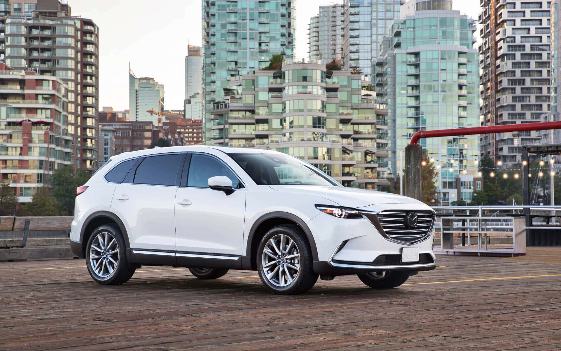 Mazda Cx-9 桌布 壁紙 — 一架時尚白色SUV停泊在木製碼頭上，背後有現代玻璃摩天大樓聳立。