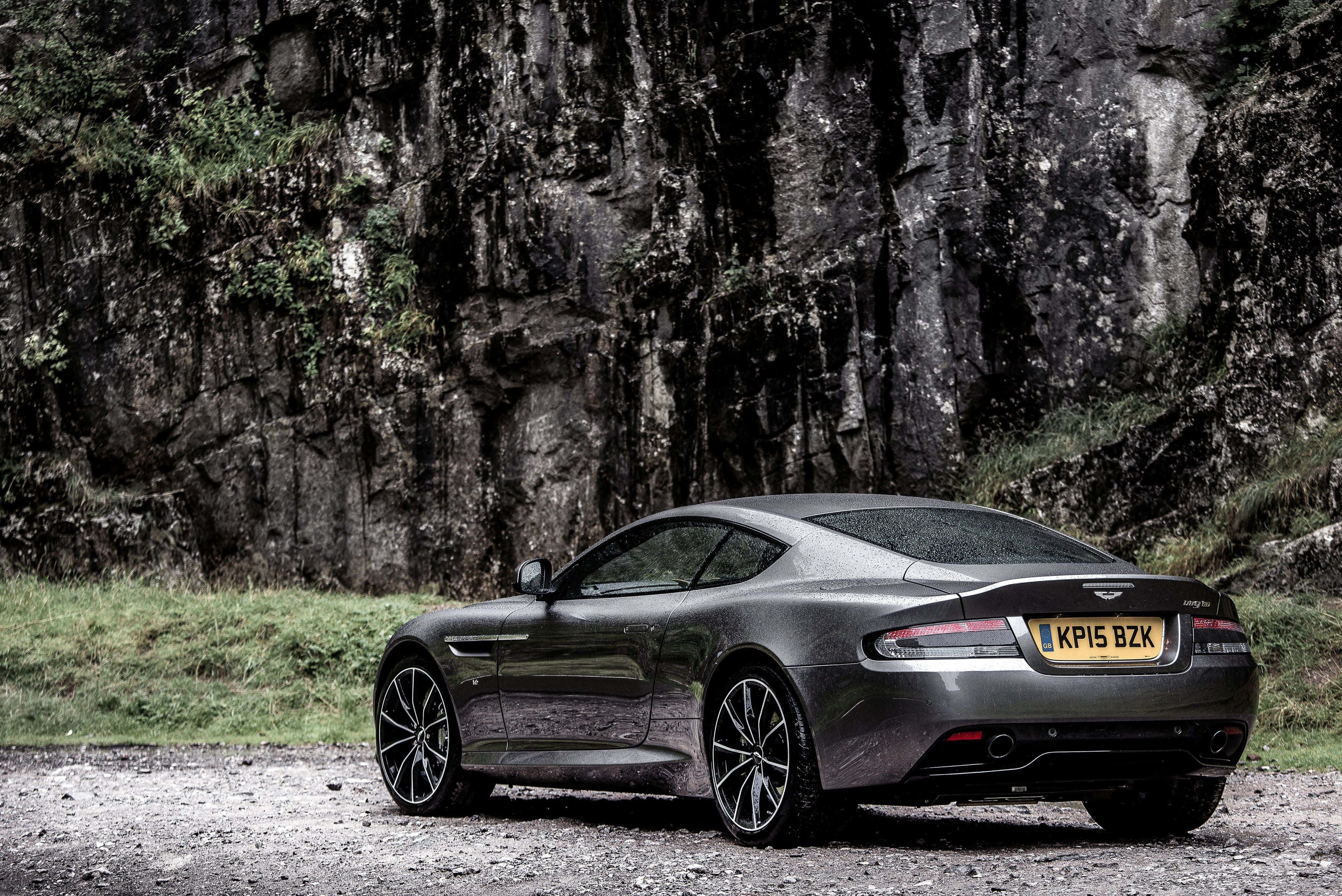 Aston Martin Db9 桌布 壁紙 — 一架時尚的啞光灰色 Aston Martin 跑車停泊在佈滿青苔的陡峭懸崖旁邊的碎石路上。