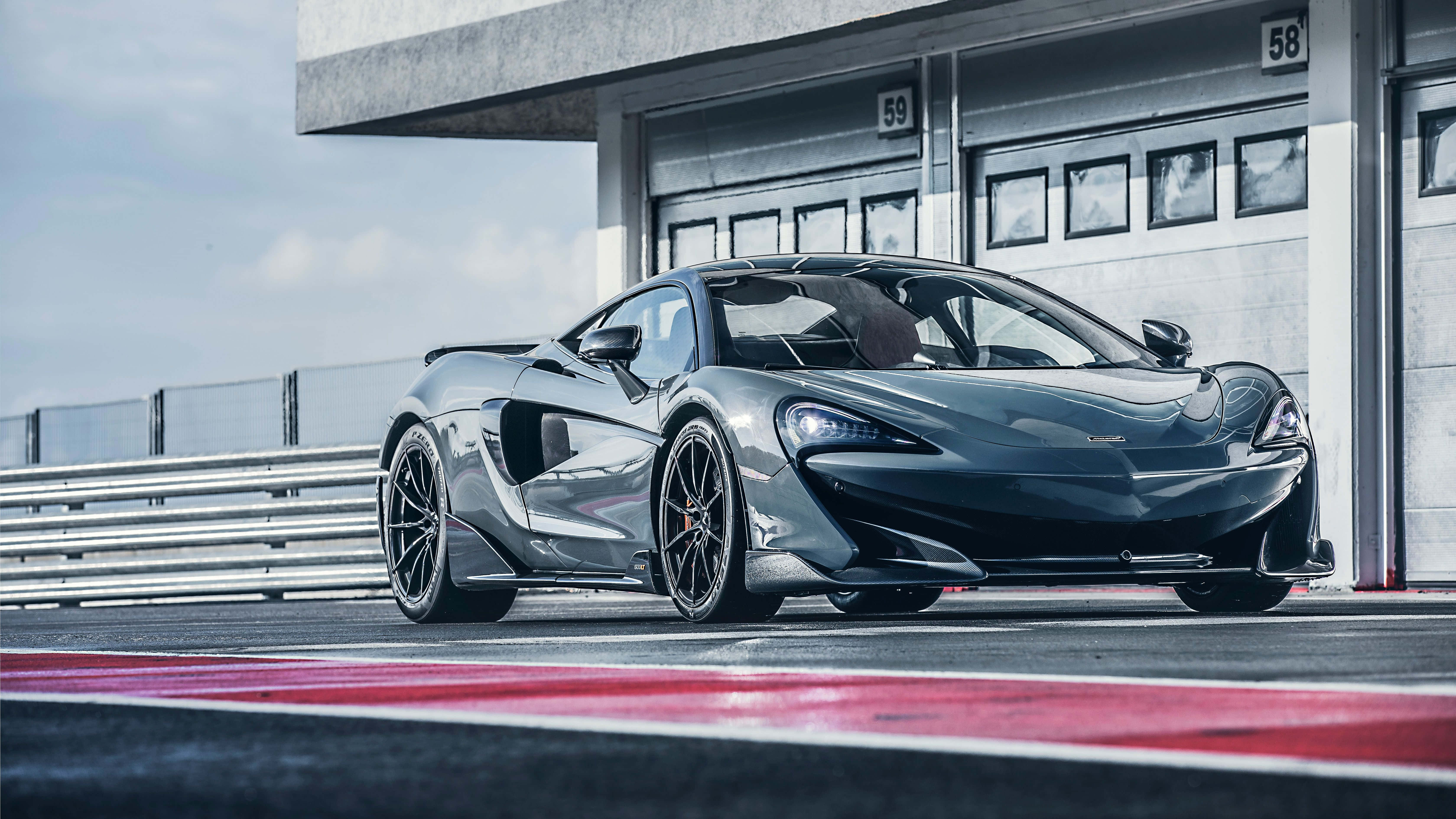 Mclaren 600lt 桌布 壁紙 — 一架時尚的啞光灰色McLaren跑車停泊在賽道上，旁邊是帶有紅色路緣石的混凝土車庫，天空晴朗。