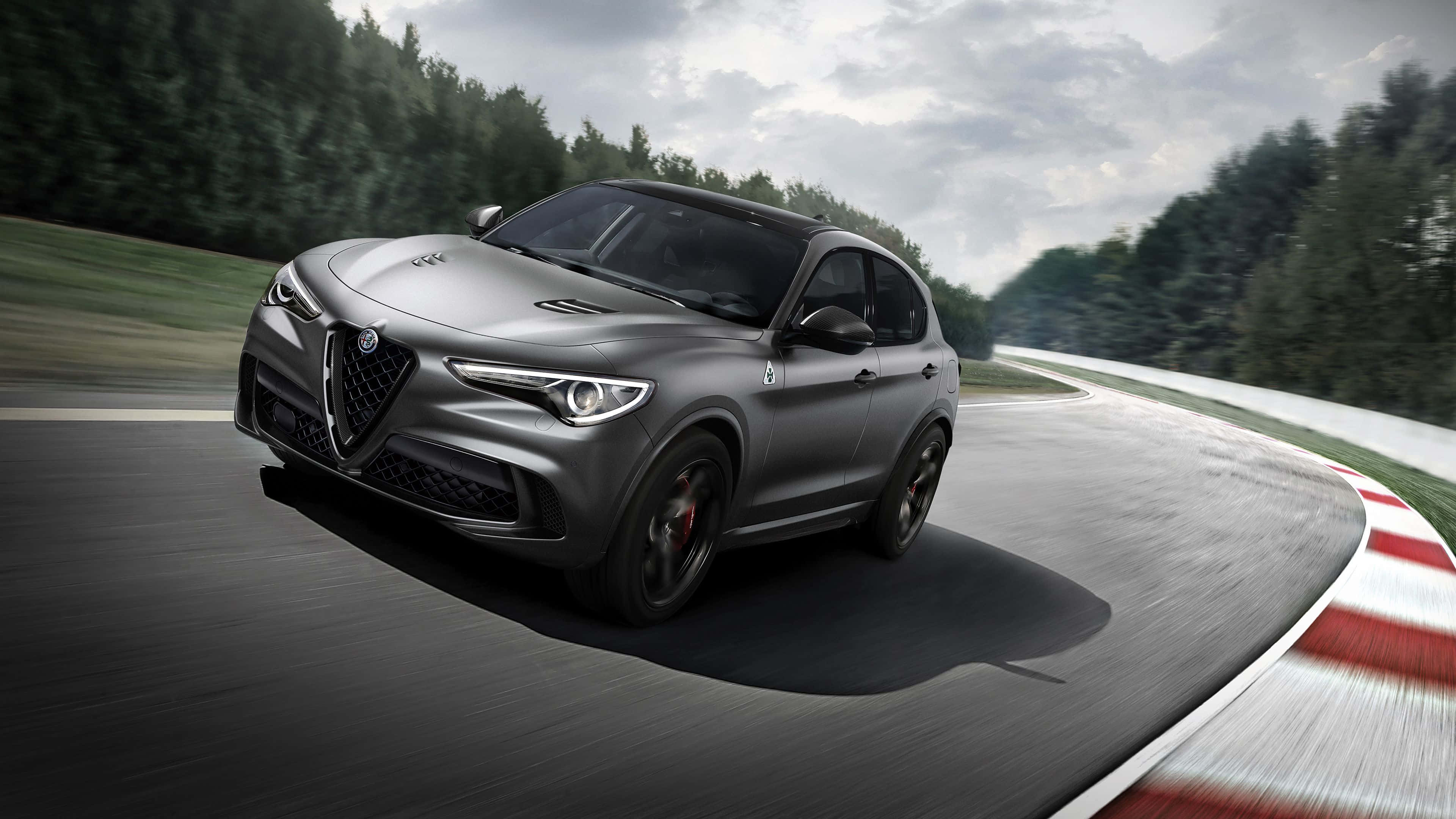 Alfa Romeo Stelvio 桌布 壁紙 — 一架時尚的啞光灰色愛快羅密歐SUV在陰天下一條彎曲的賽道上飛馳，賽道兩旁有紅白路緣和茂密的綠色森林。
