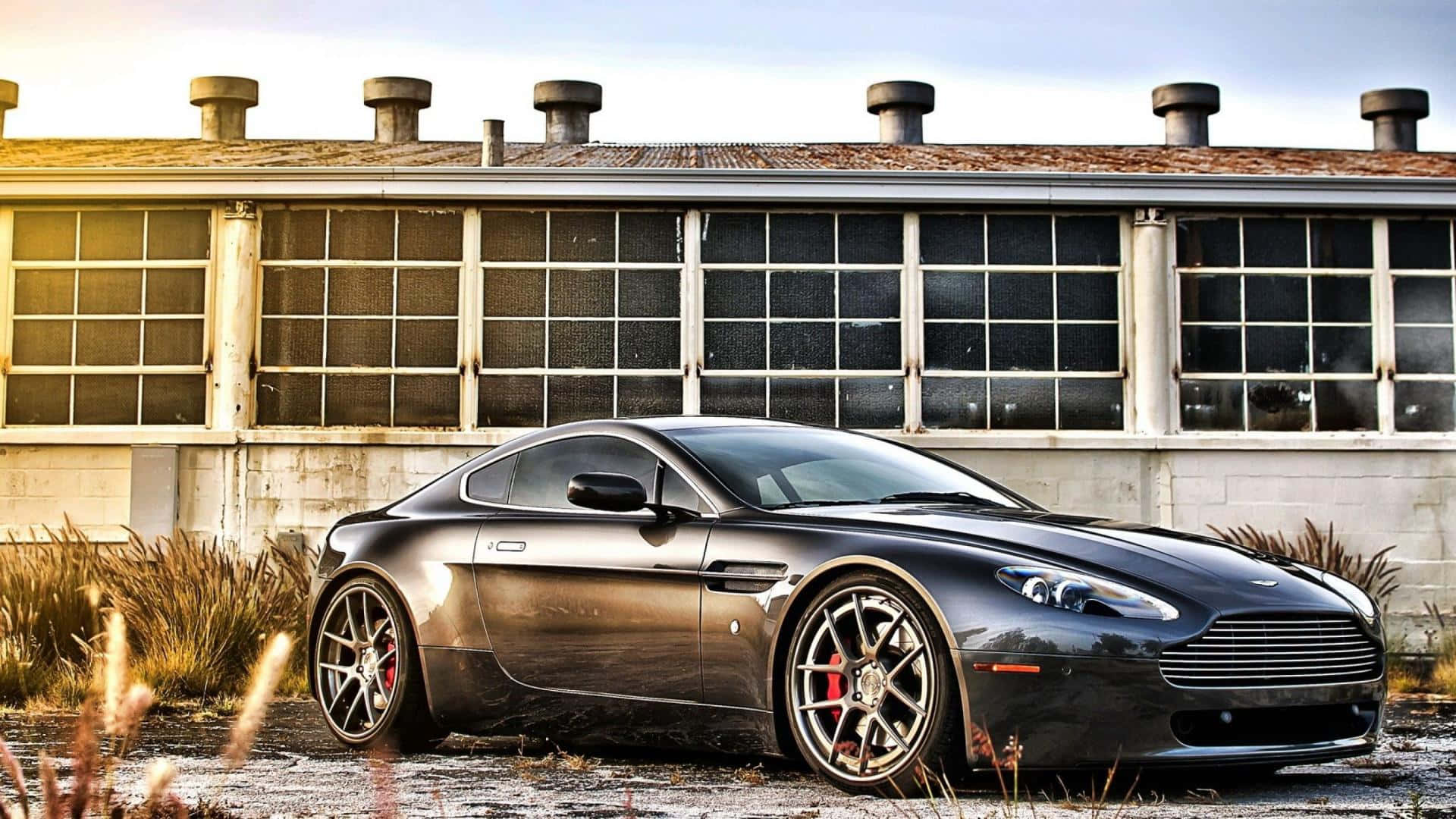 Aston Martin V12 Vantage 桌布 壁紙 — 一架時尚的啞光黑色Aston Martin跑車停泊在一個帶有大窗戶、風化了的工業建築前。