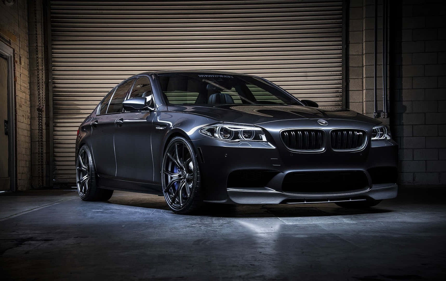 Bmw M5 桌布 壁紙 — 一架時尚的啞光黑色BMW M5運動轎車停泊在光線昏暗的工業車庫中，車庫有混凝土地板和金屬捲門。