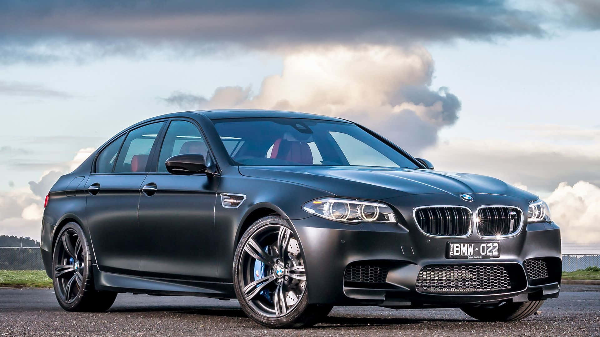 Bmw M5 桌布 壁紙 — 一架時尚的啞光黑色BMW M5運動轎車停在瀝青路上，背景是戲劇性的多雲天空，車牌清晰可見。