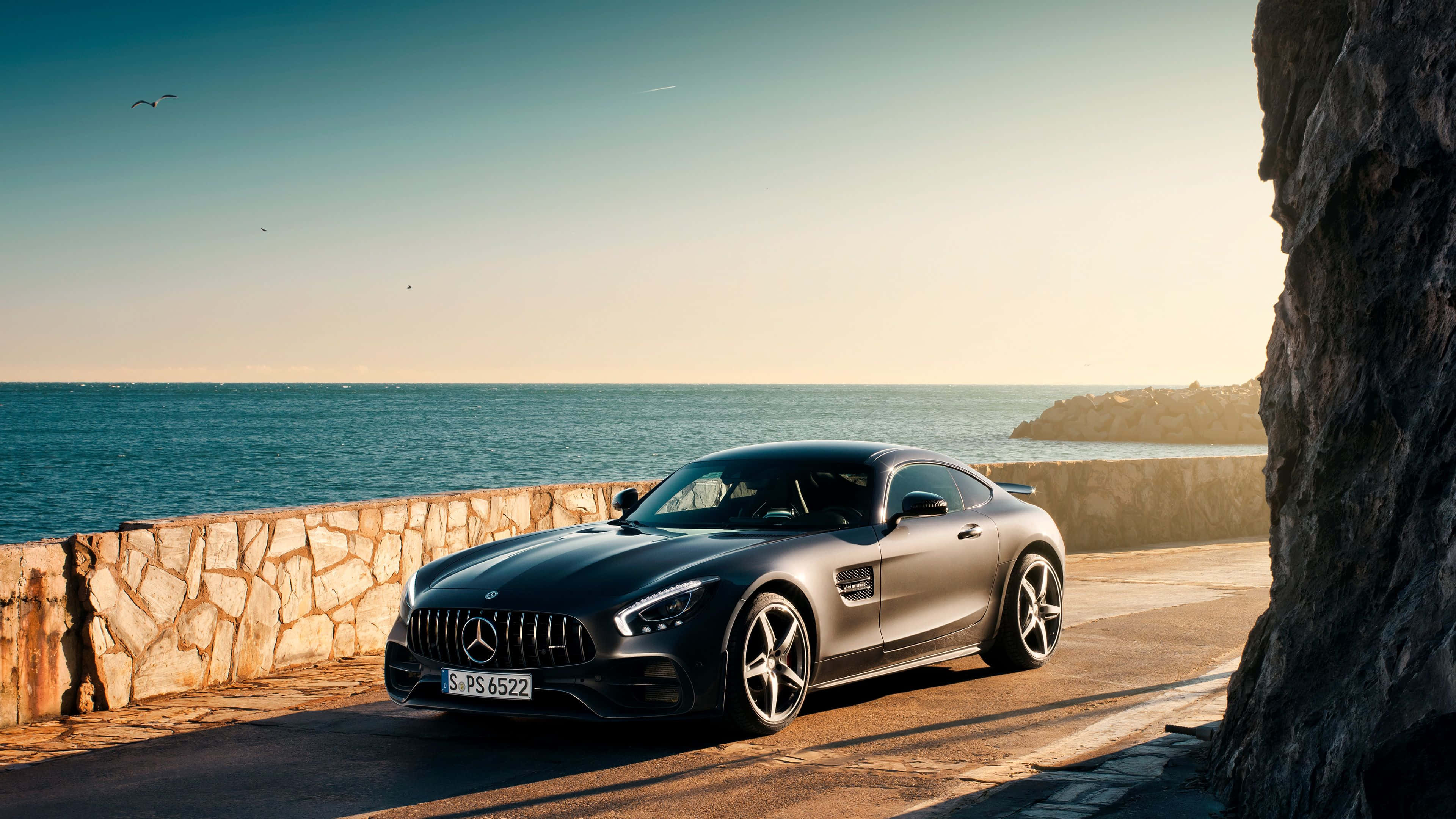 4K Mercedes 桌布 壁紙 — 一架時尚的啞光黑色Mercedes-AMG GT跑車停泊在沿海道路上，旁邊是石牆，背景是日落時分的海洋和懸崖。