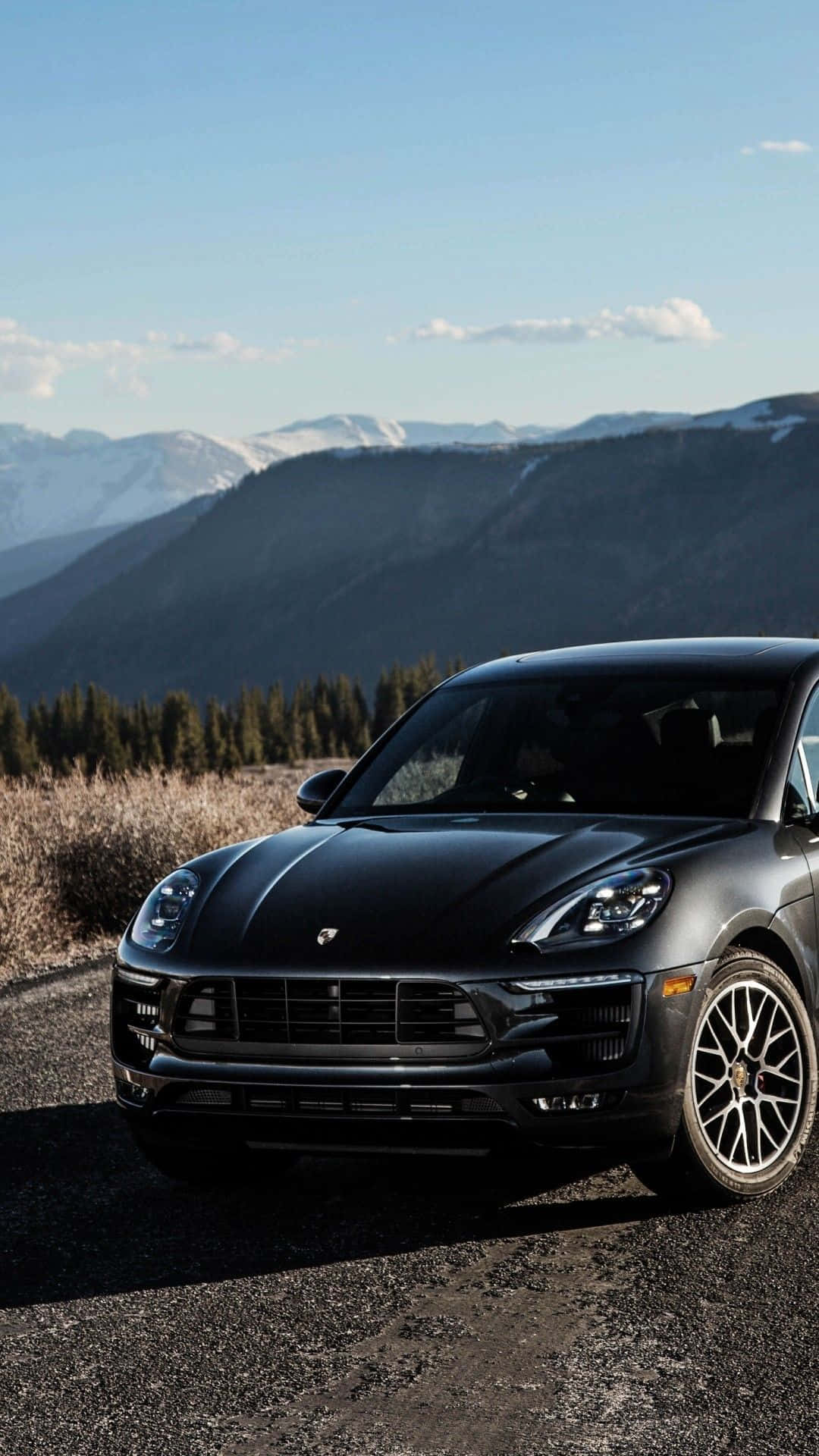 Porsche iPhone 桌布 壁紙 — 一架時尚的啞光黑色保時捷 Macan 停在碎石路上，背景是白雪覆蓋的山脈和常青樹，天空清澈湛藍。